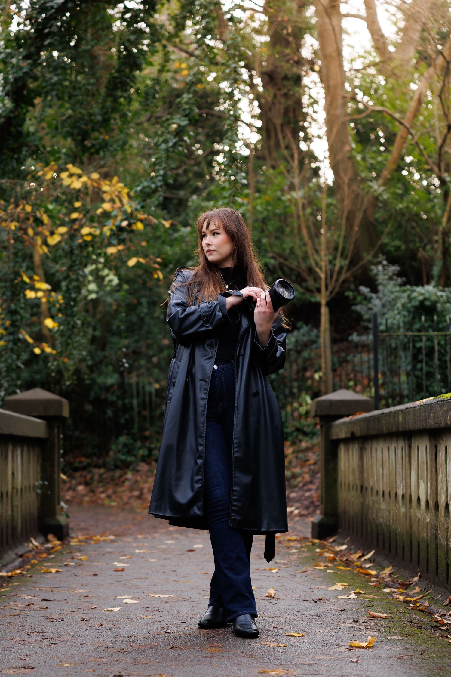 A figure in a dark coat walks along a wooden fence-lined path covered in autumn leaves on a cloudy day.