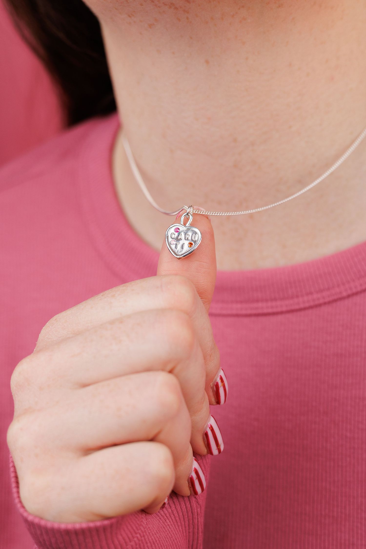 Close-up shots of hands displaying a delicate silver heart necklace against a pink background.