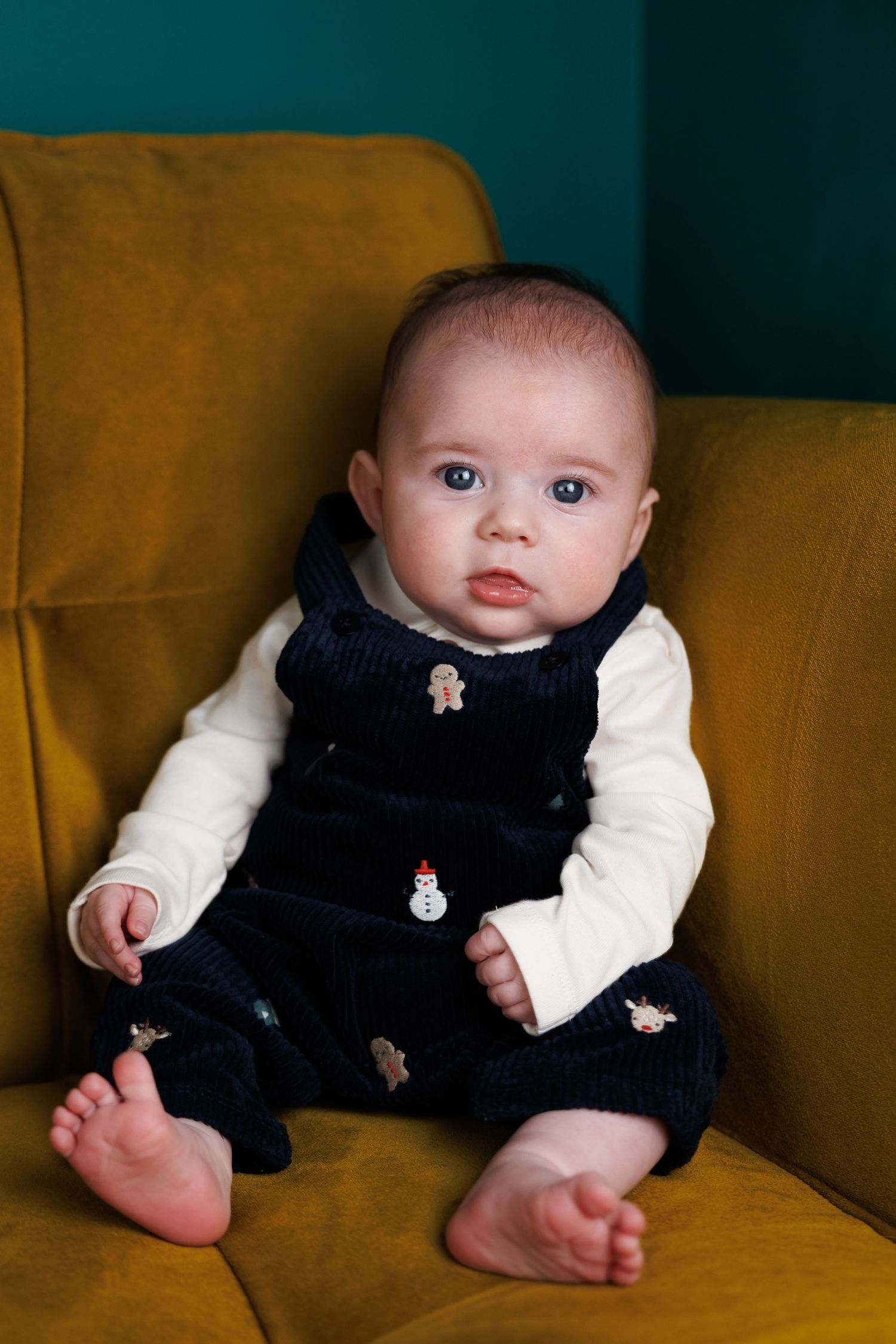 Baby wearing white long sleeve shirt and navy overalls with embroidered details sits on mustard yellow couch.