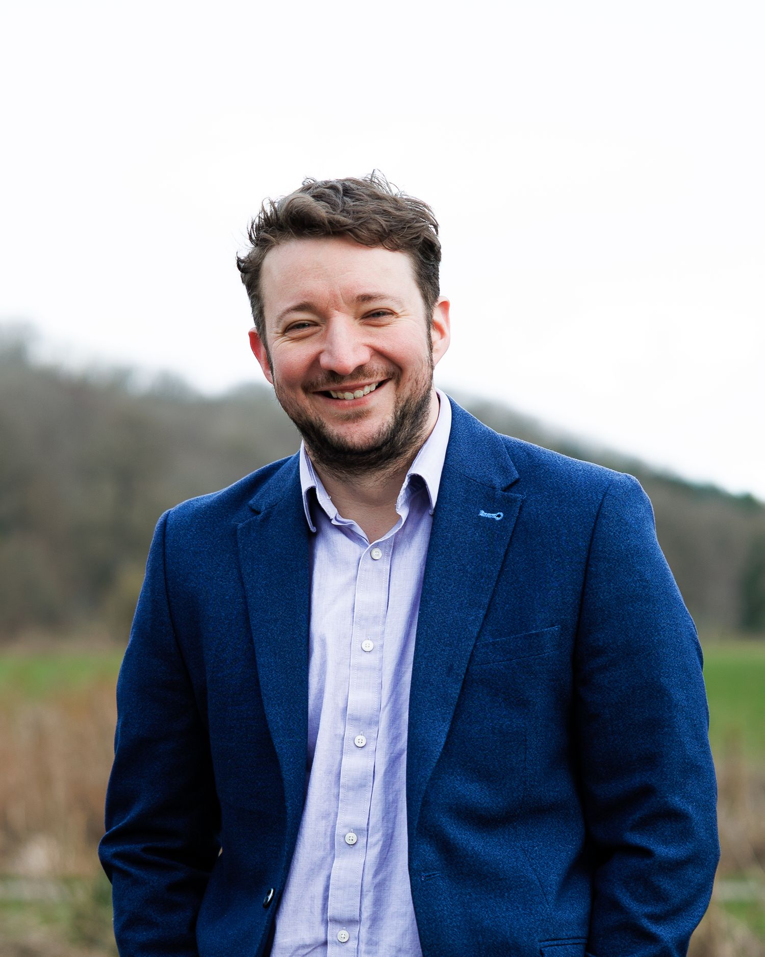 Professional in a navy blazer and white shirt smiles warmly against a blurred outdoor background.