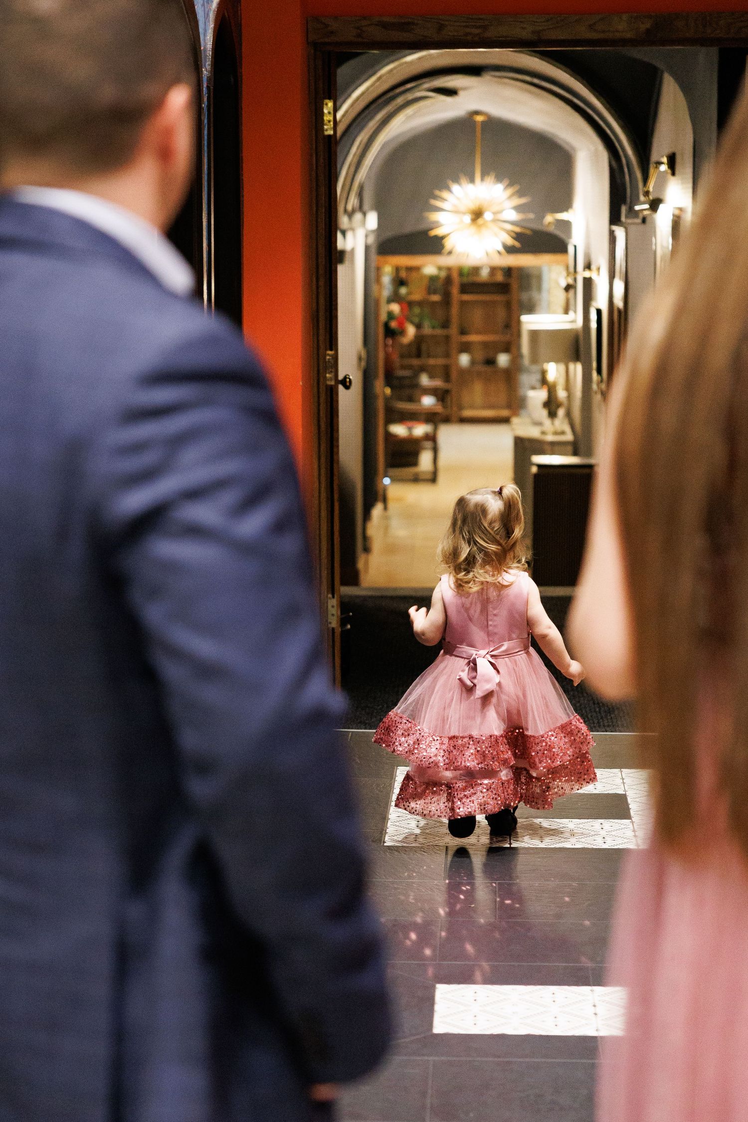 A small child in a pink tulle dress twirls playfully in front of a mirror while adults watch in the background.
