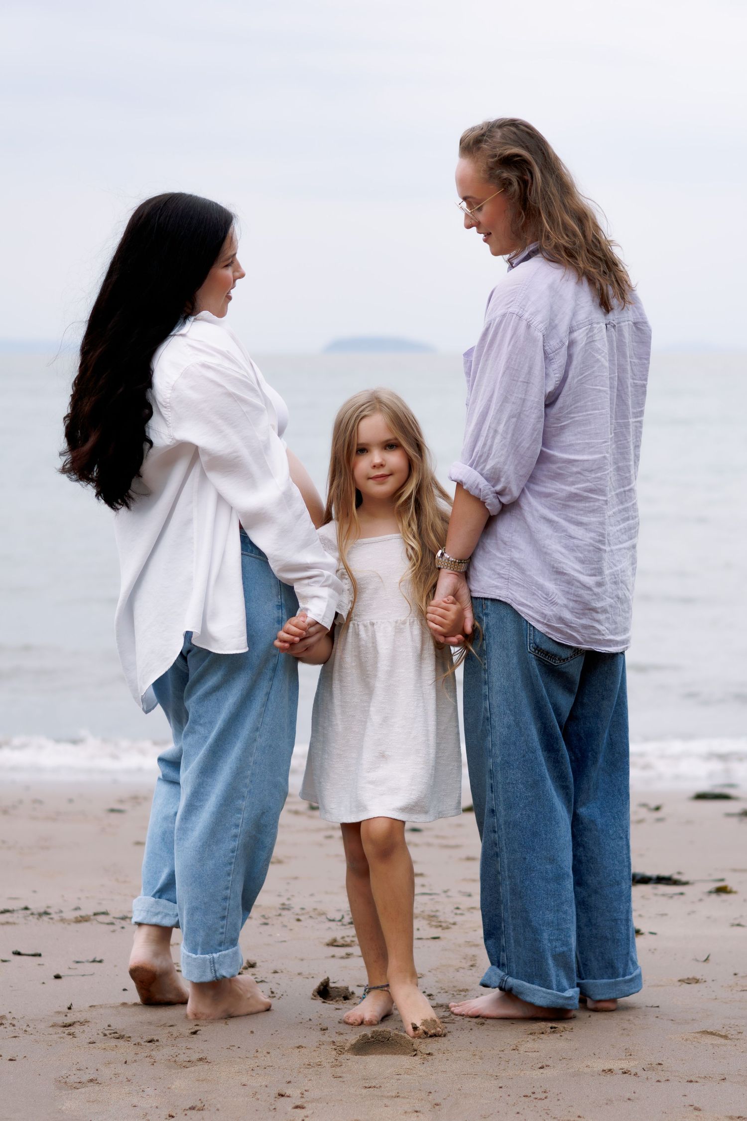 Family trio walking on a sandy beach wearing casual denim and white outfits.