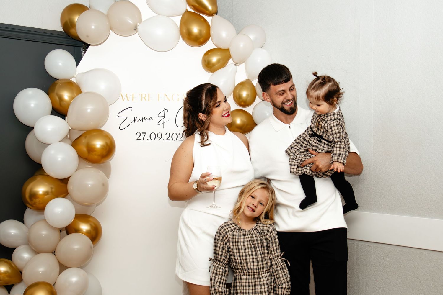 A balloon arch with gold and white balloons decorates a white wall during a celebration event.