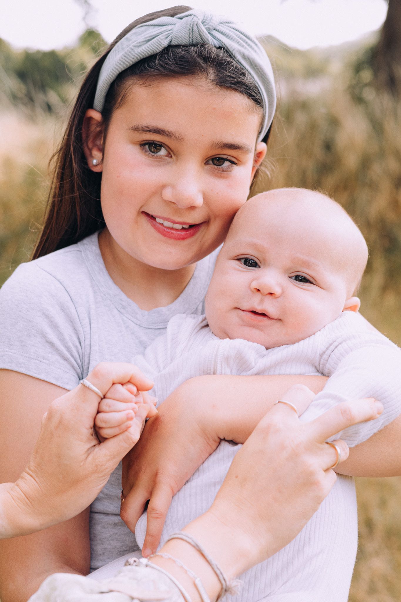 A sweet outdoor photoshoot shows a young girl in grey holding and interacting with a baby in natural lighting.