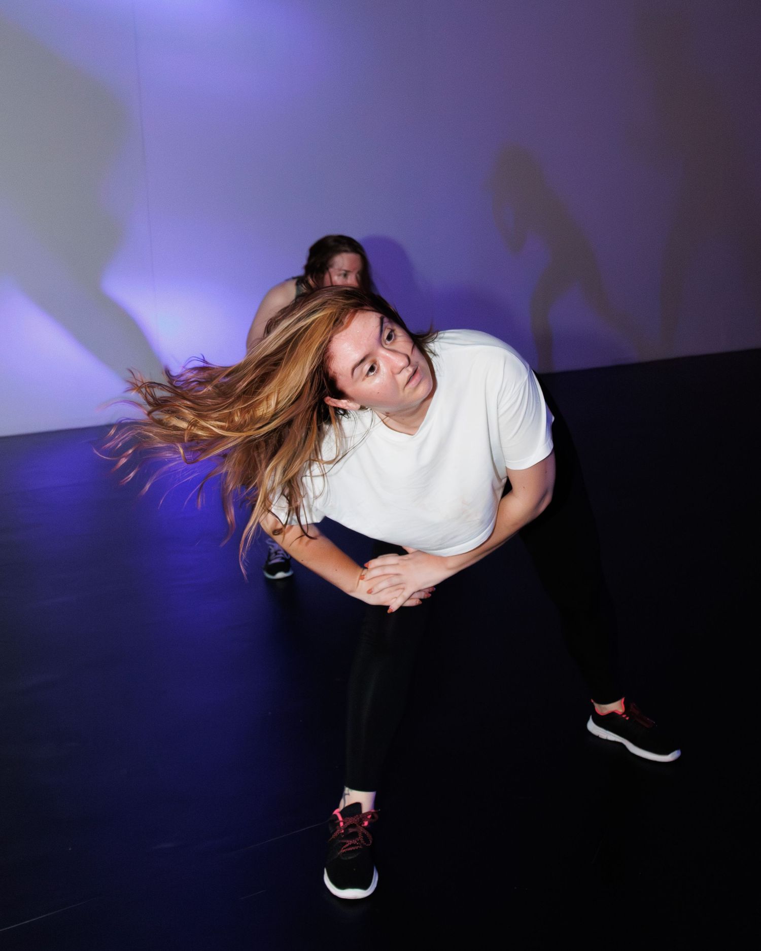 Person with long hair dancing energetically against a purple-lit wall in casual white t-shirt and black pants.
