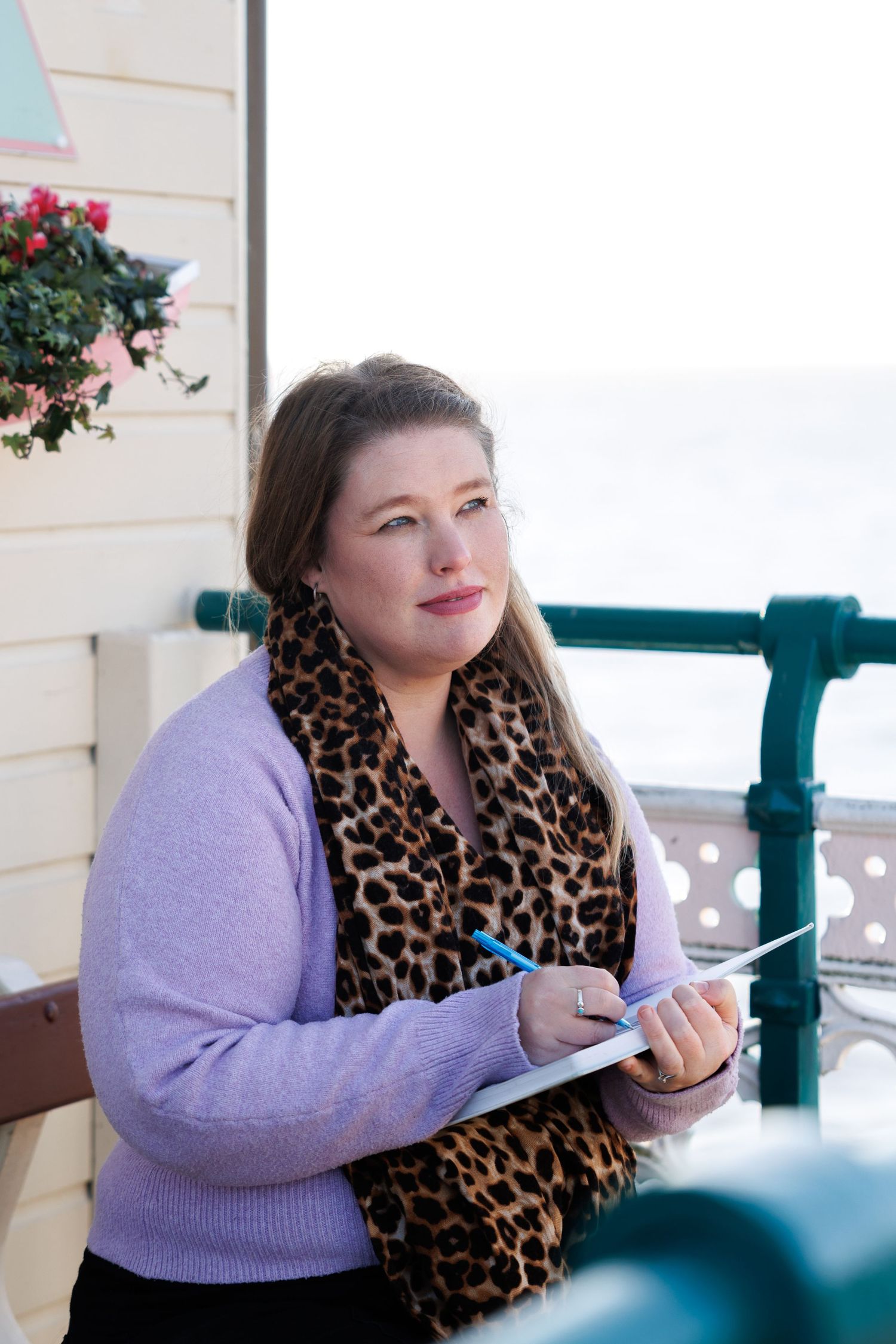 Person in purple sweater and leopard print scarf sits at table with notebook and pen.
