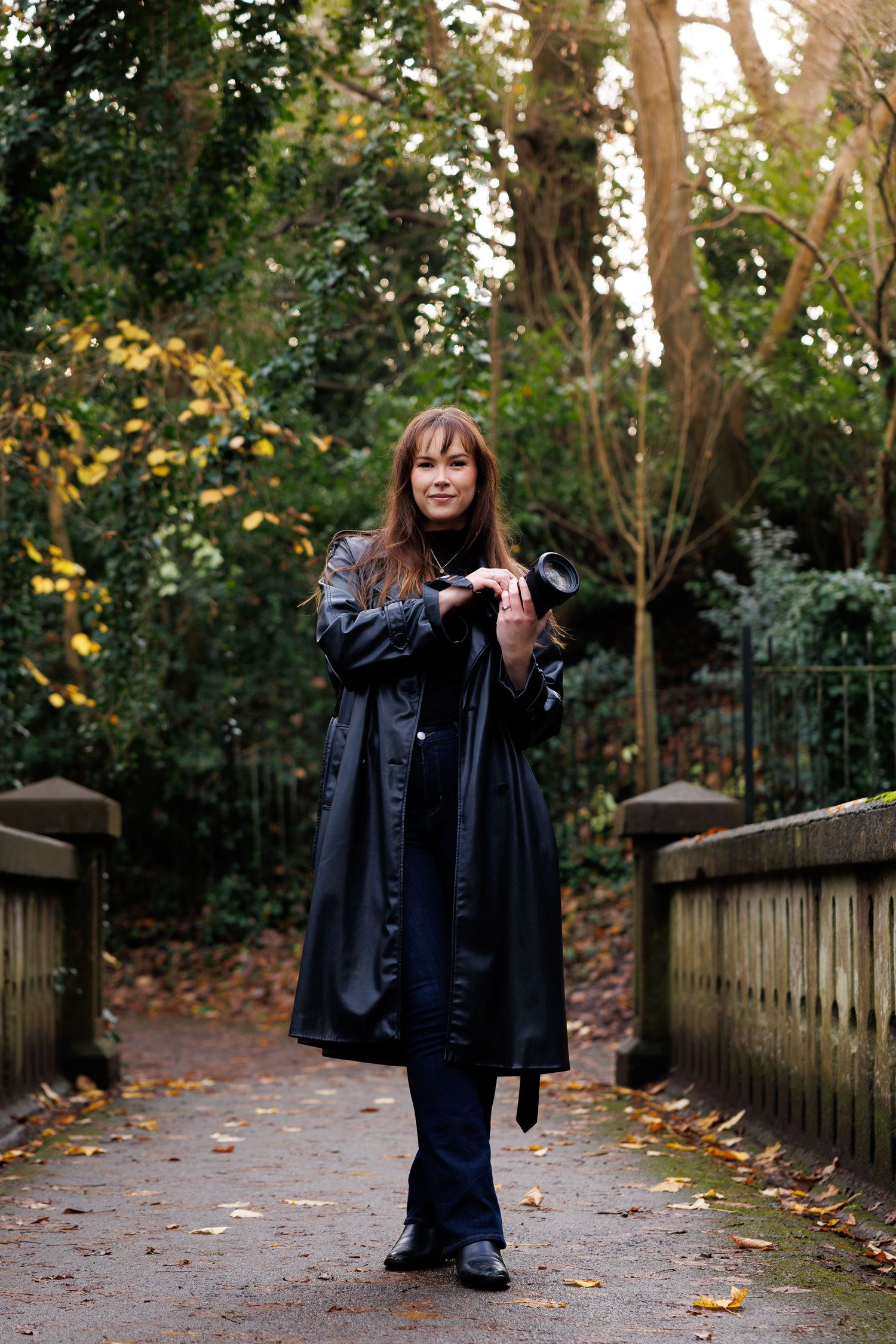 A person wearing a black coat poses playfully on a wooden bridge pathway surrounded by autumn trees.