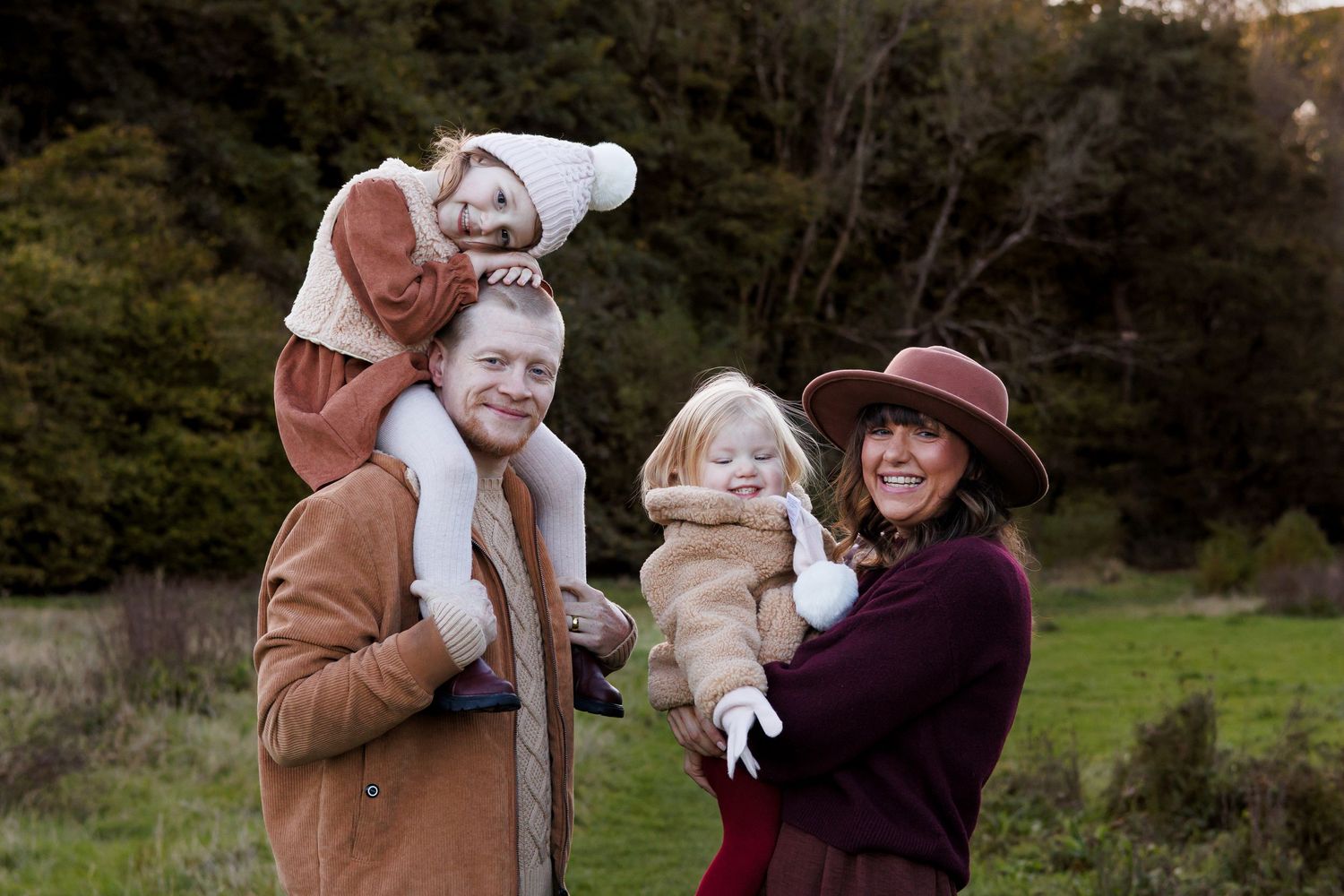 A family wearing warm autumn clothing poses together outdoors in a natural setting at sunset.