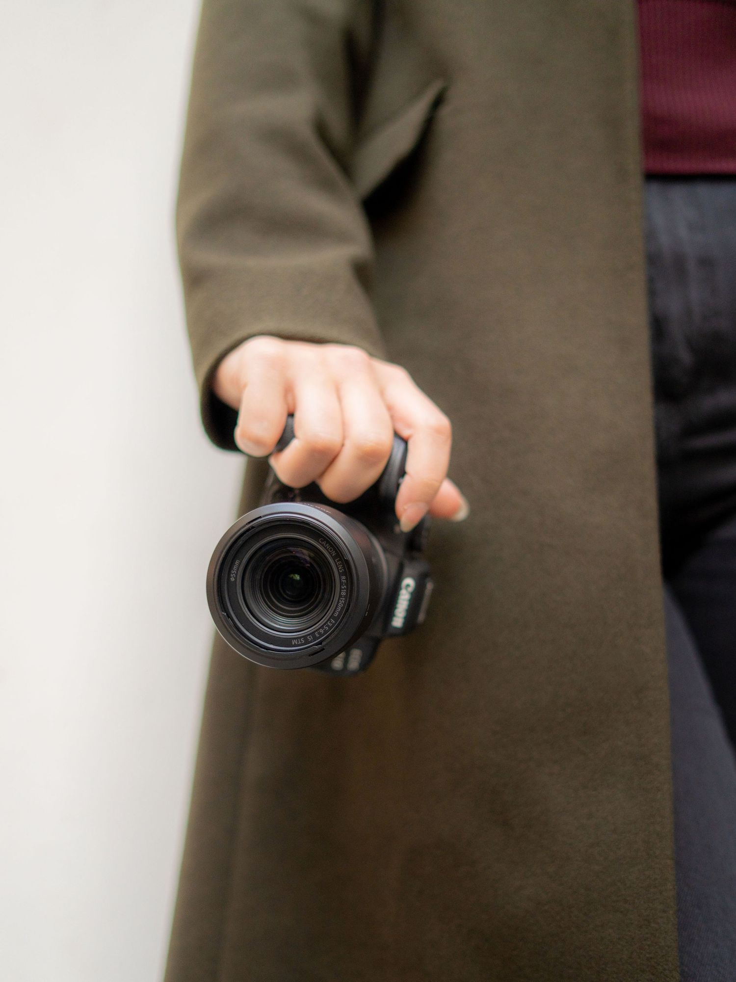 A hand in a brown coat sleeve holds a black camera lens against a light background.
