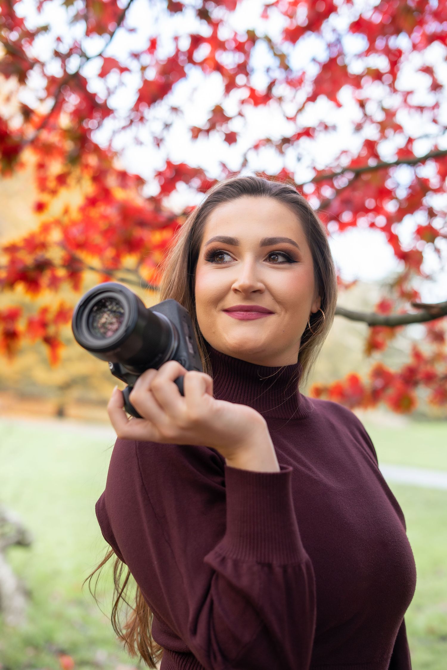 Photographer in burgundy sweater captures autumn photos beneath vibrant red maple leaves.