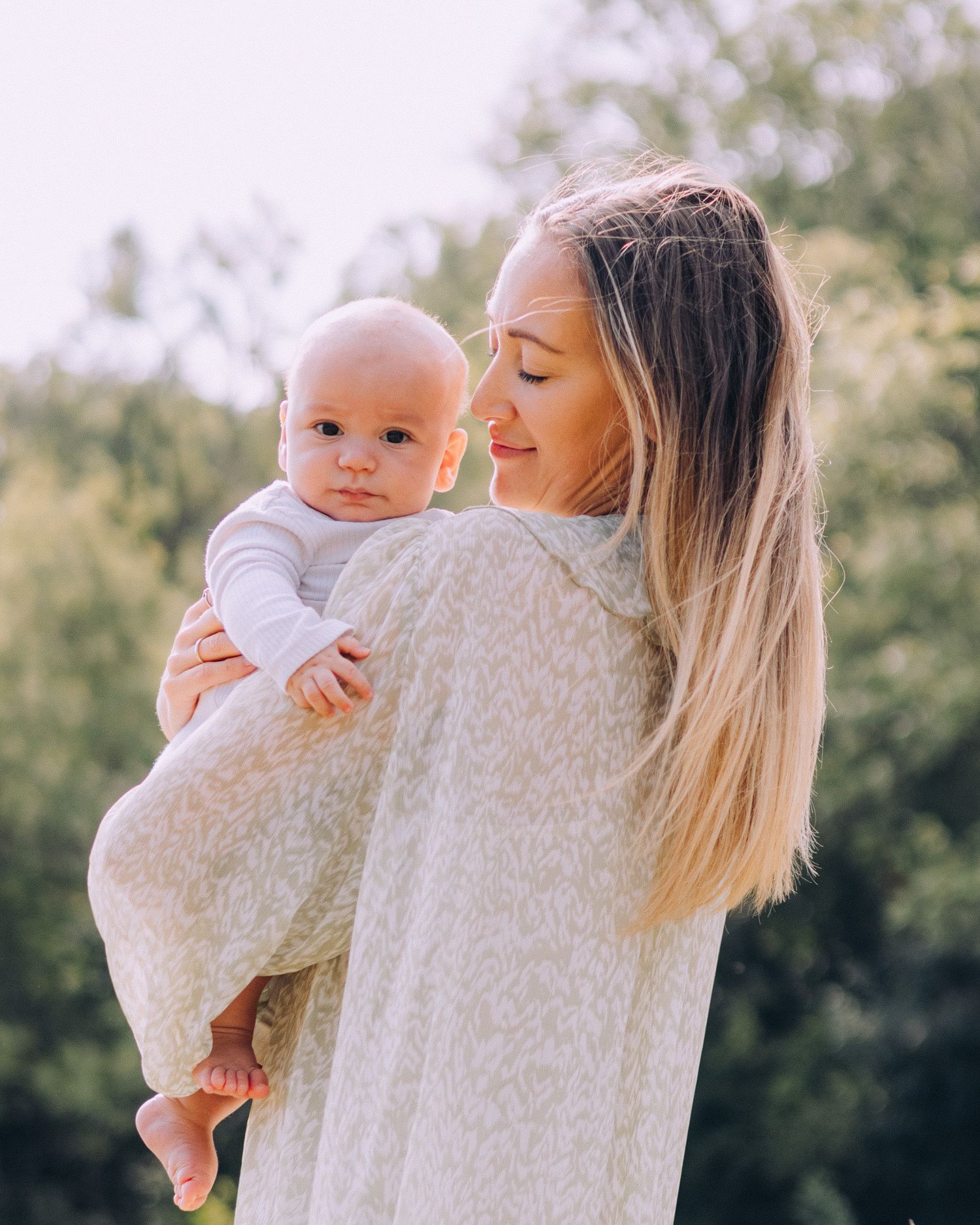 A parent in a cream sweater holds a baby in white clothing outdoors with greenery in the background.