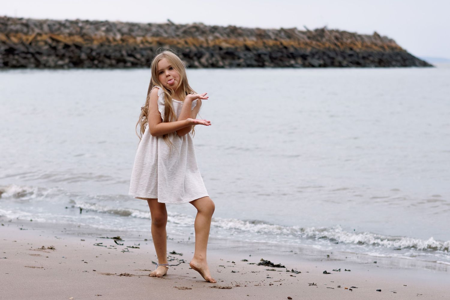 A person in a white dress walks along a sandy beach near a rocky shoreline.