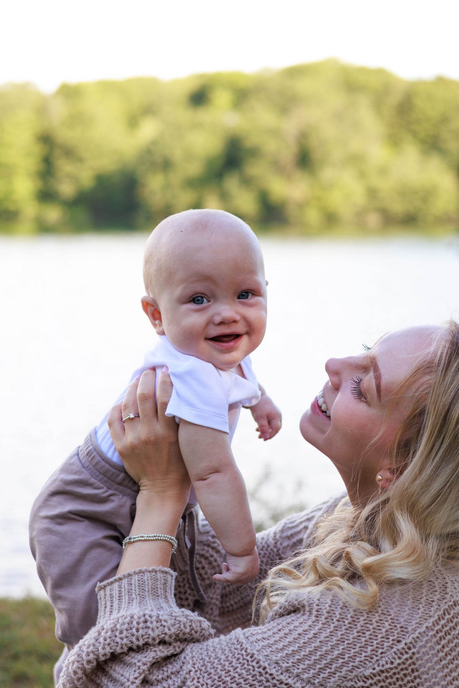 A parent in a knit sweater lifts and plays with a baby wearing white against an outdoor backdrop near a lake.
