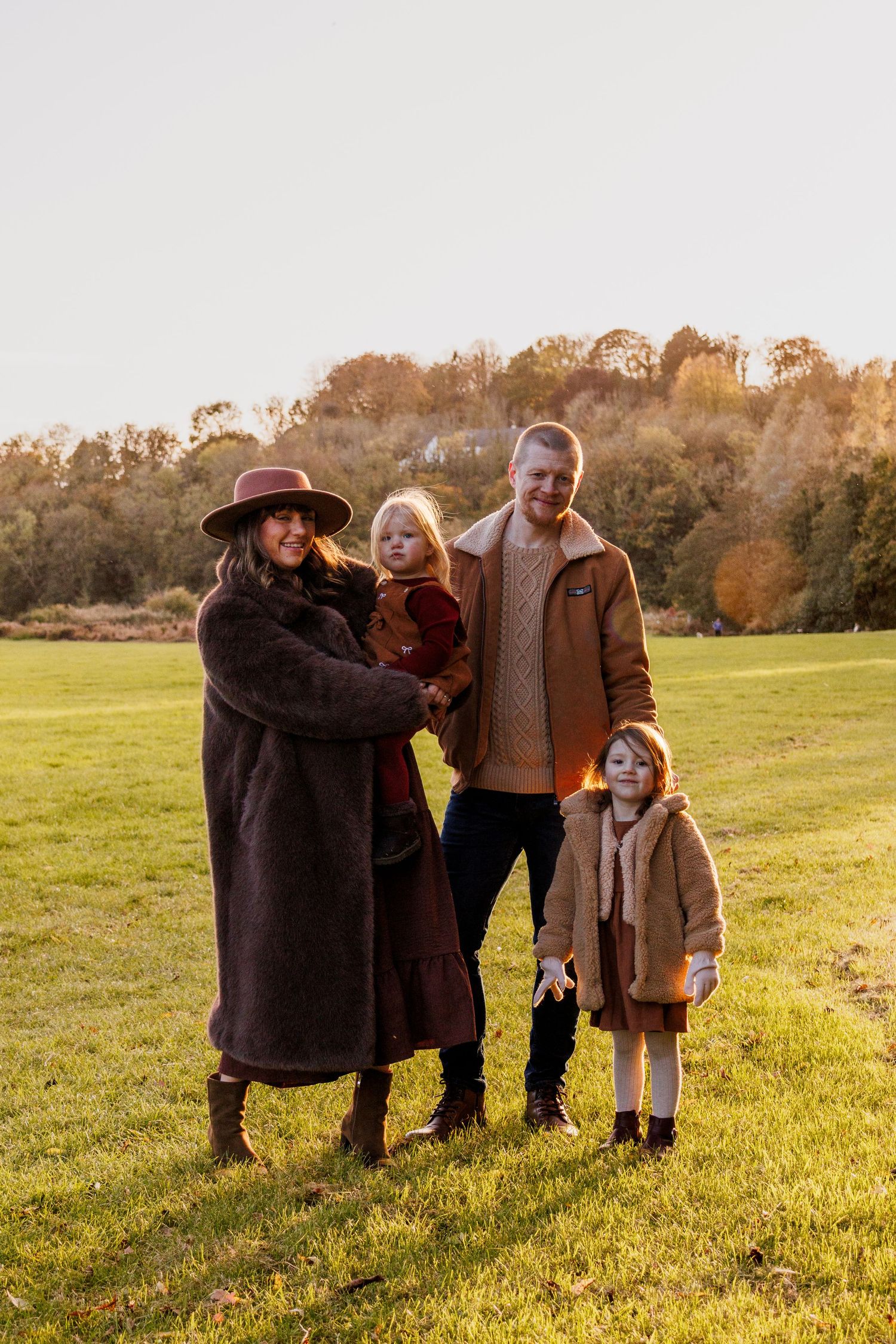 A family stands together in a grassy field at sunset wearing cozy fall outfits in warm brown tones.