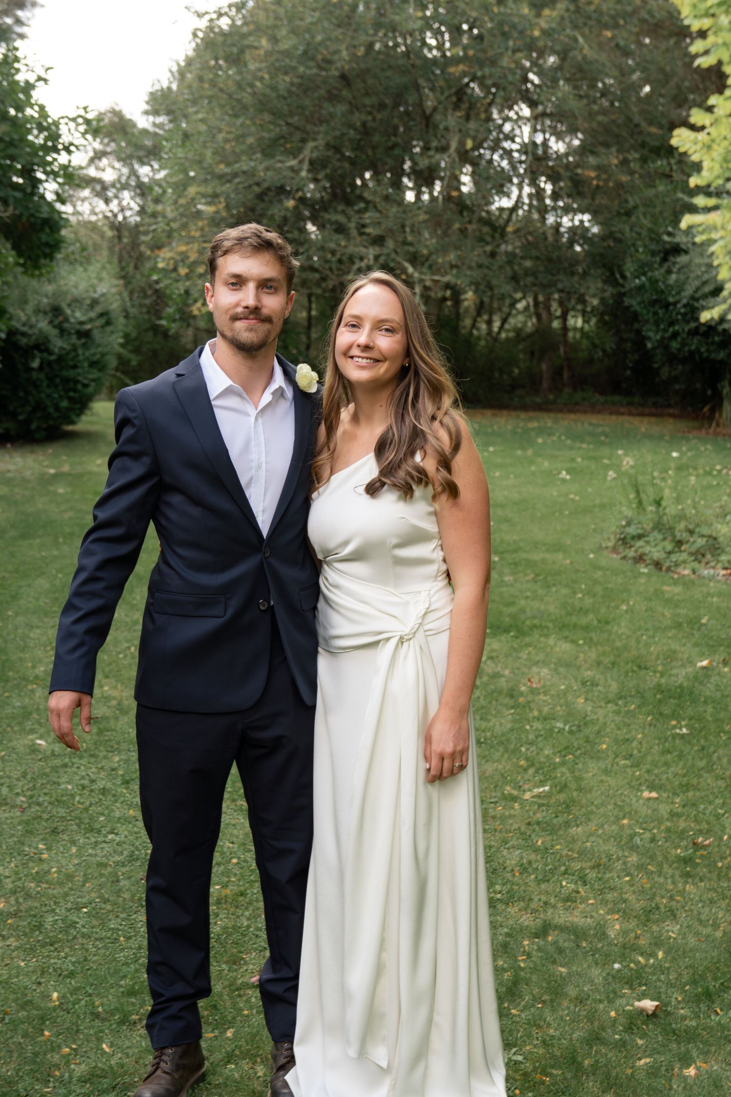 A smiling bride in a white gown and groom in a navy suit pose together outdoors on a green lawn.