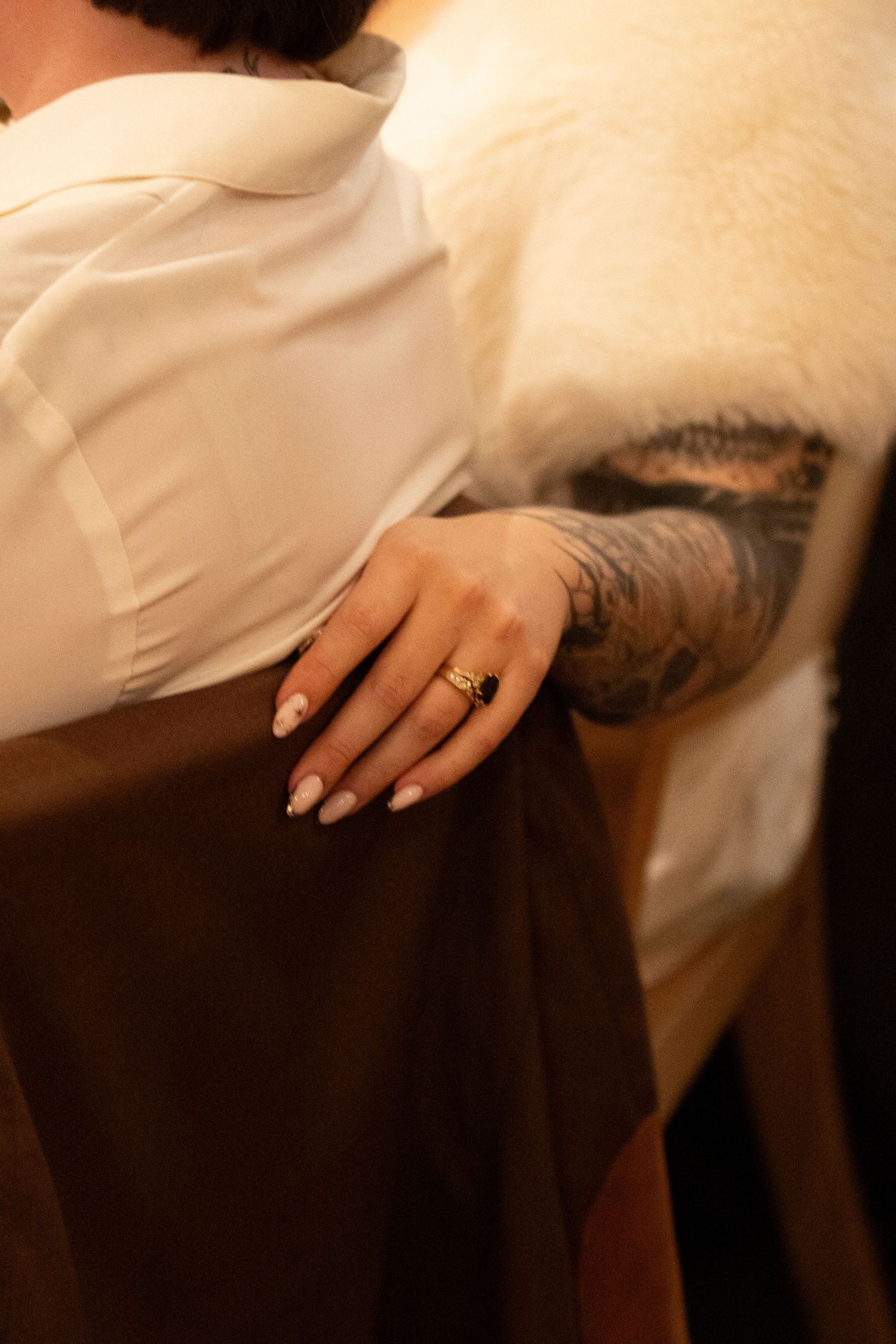 Close-up of a hand with nude nails and gold ring resting on brown skirt, beside a heavily tattooed arm.