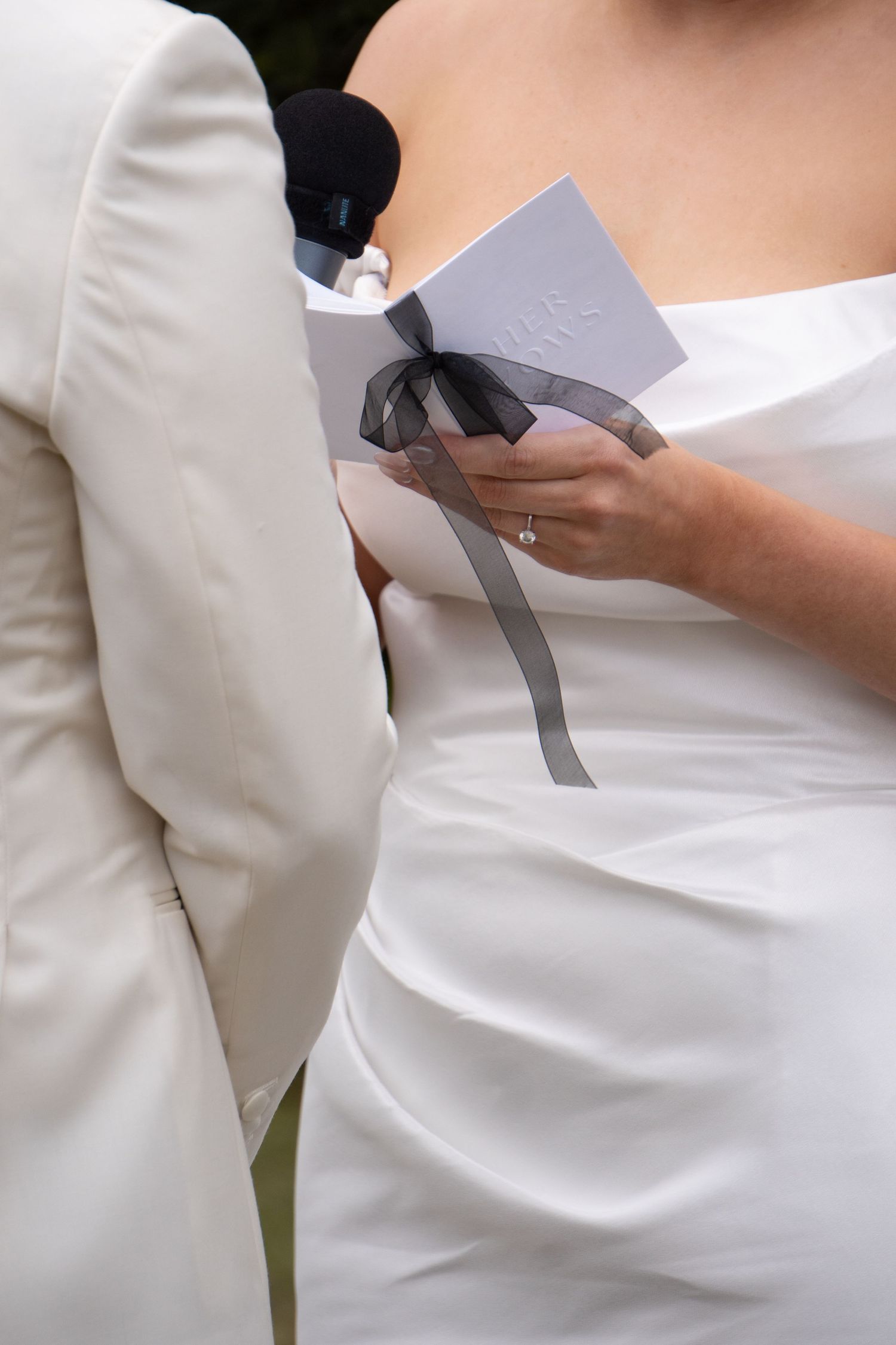 Bride in white strapless gown holding wedding program with black ribbon bow during outdoor ceremony.