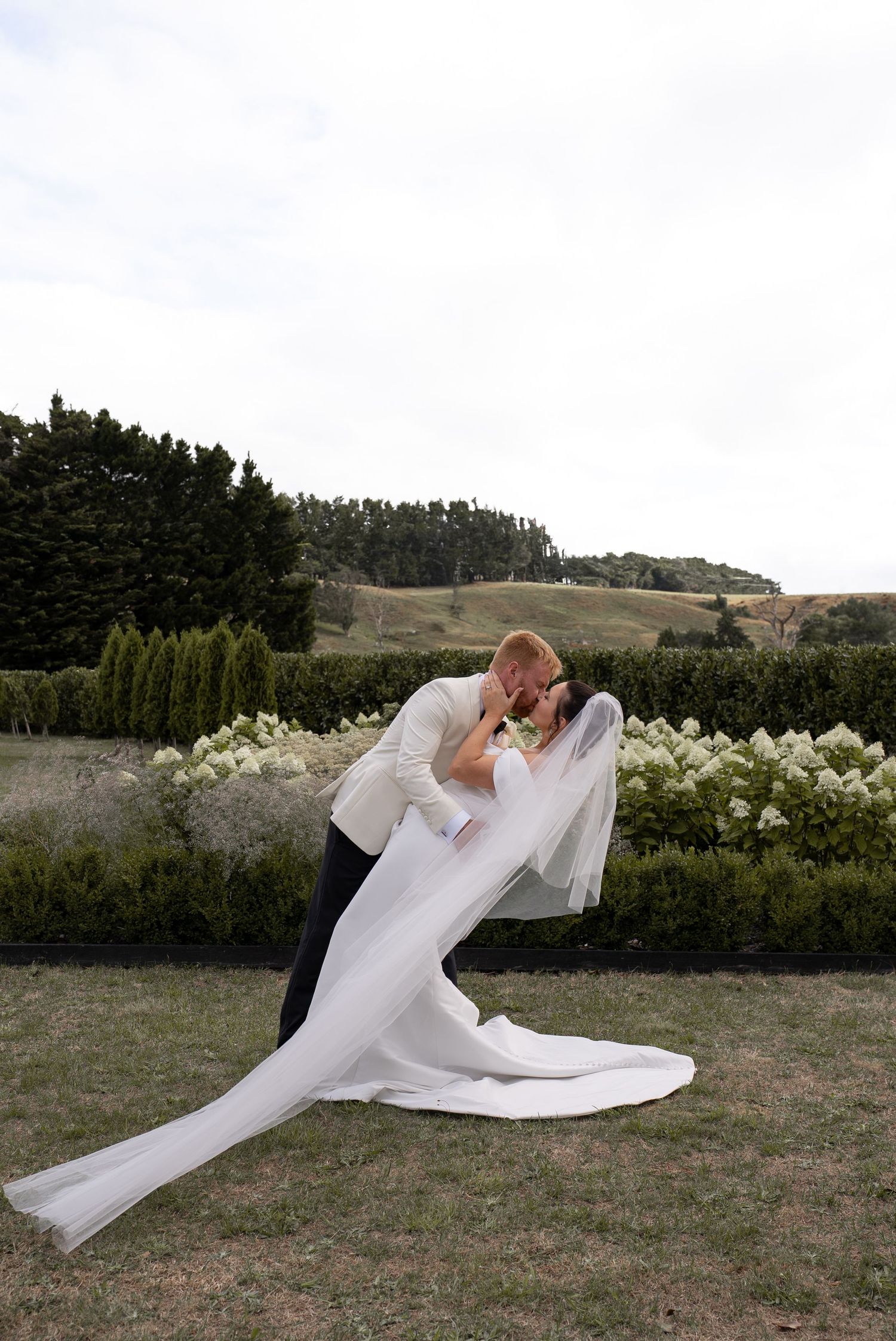 Groom dips bride in a romantic kiss outdoors, her long veil flowing, surrounded by lush greenery on their wedding day.