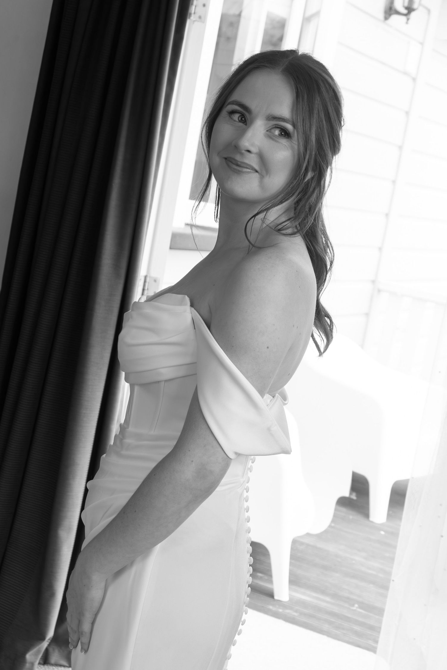 Black and white photo of a bride in an off-shoulder wedding gown, glancing back with a smile near a window.