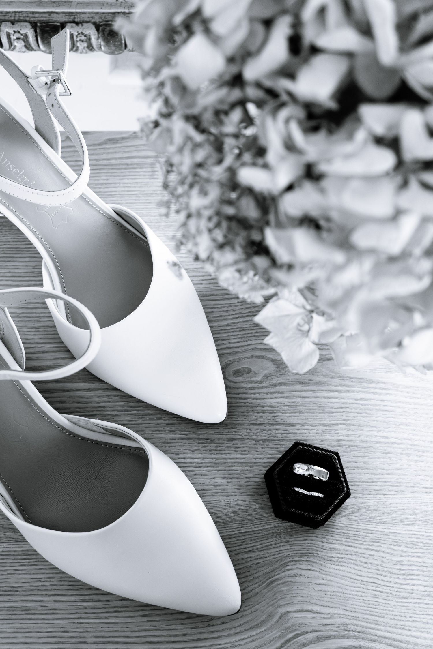 Black and white photo of white bridal heels, a diamond ring in a box, and floral bouquet on wooden surface.