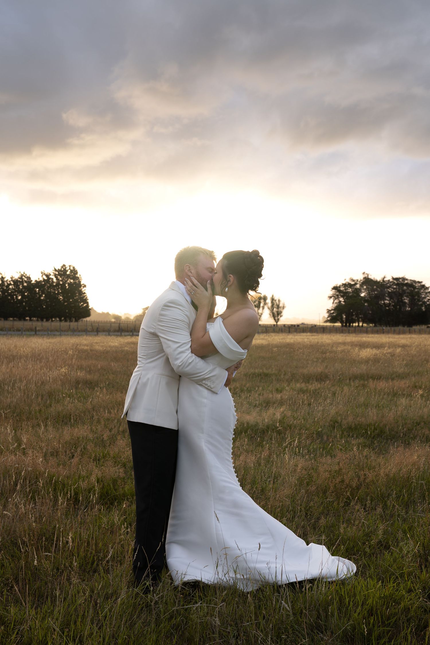 Bride and groom share a romantic kiss at sunset in an open field, bride in white gown, groom in white jacket.