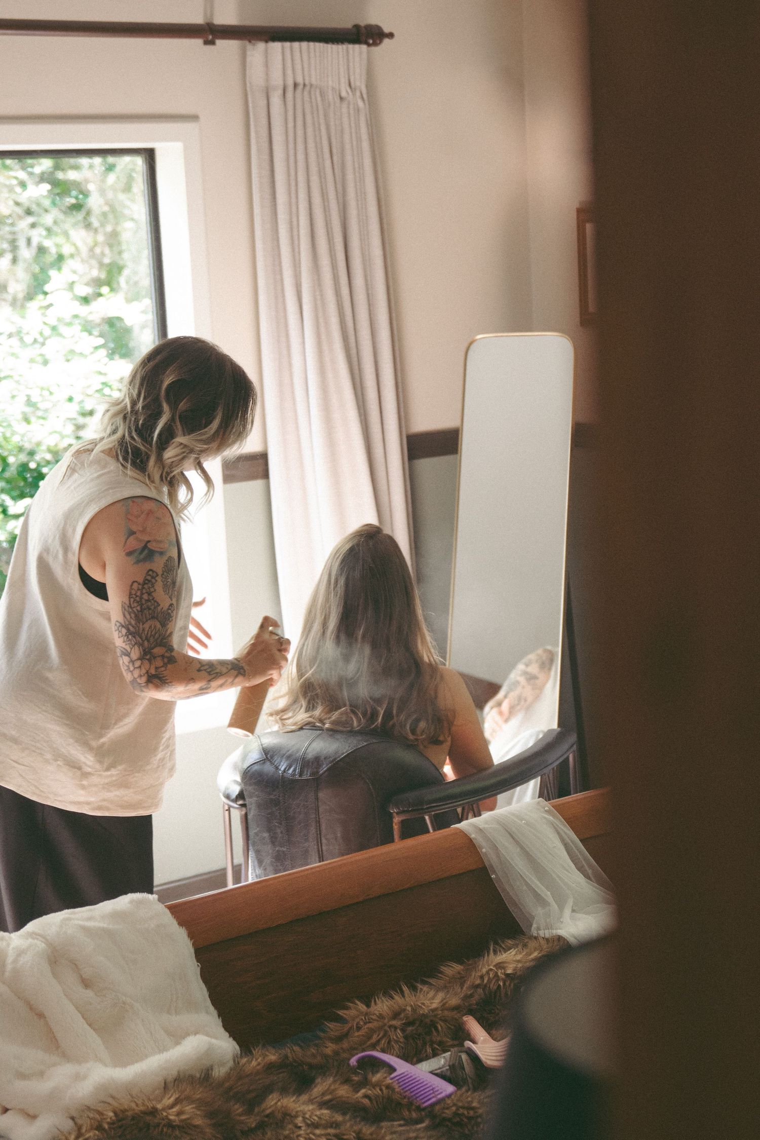 A tattooed woman styles the hair of a seated woman in front of a mirror near a bright window indoors.