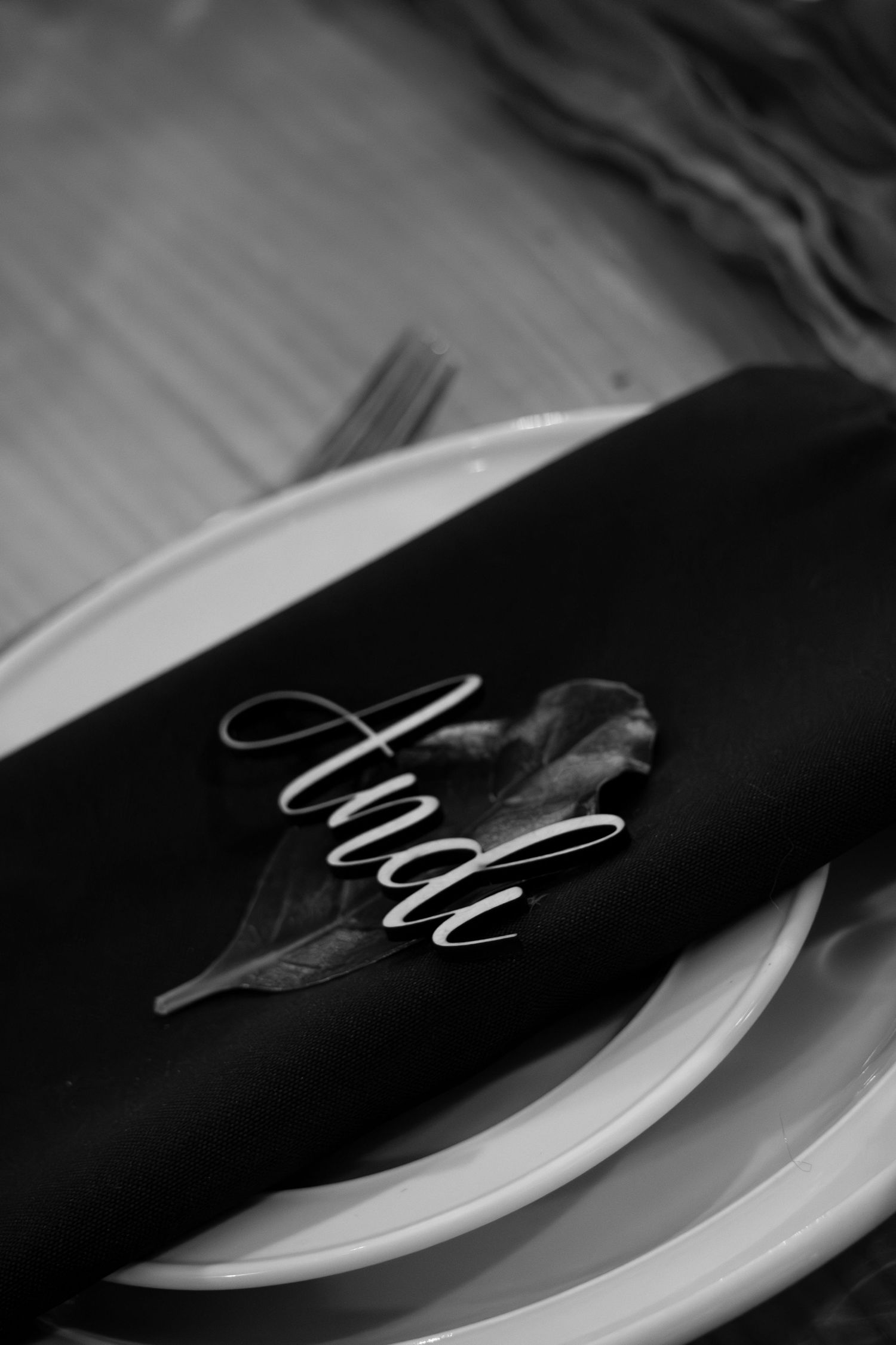 Black and white photo of elegant place setting with dark napkin, leaf, and cursive name card reading Andi on white plate.