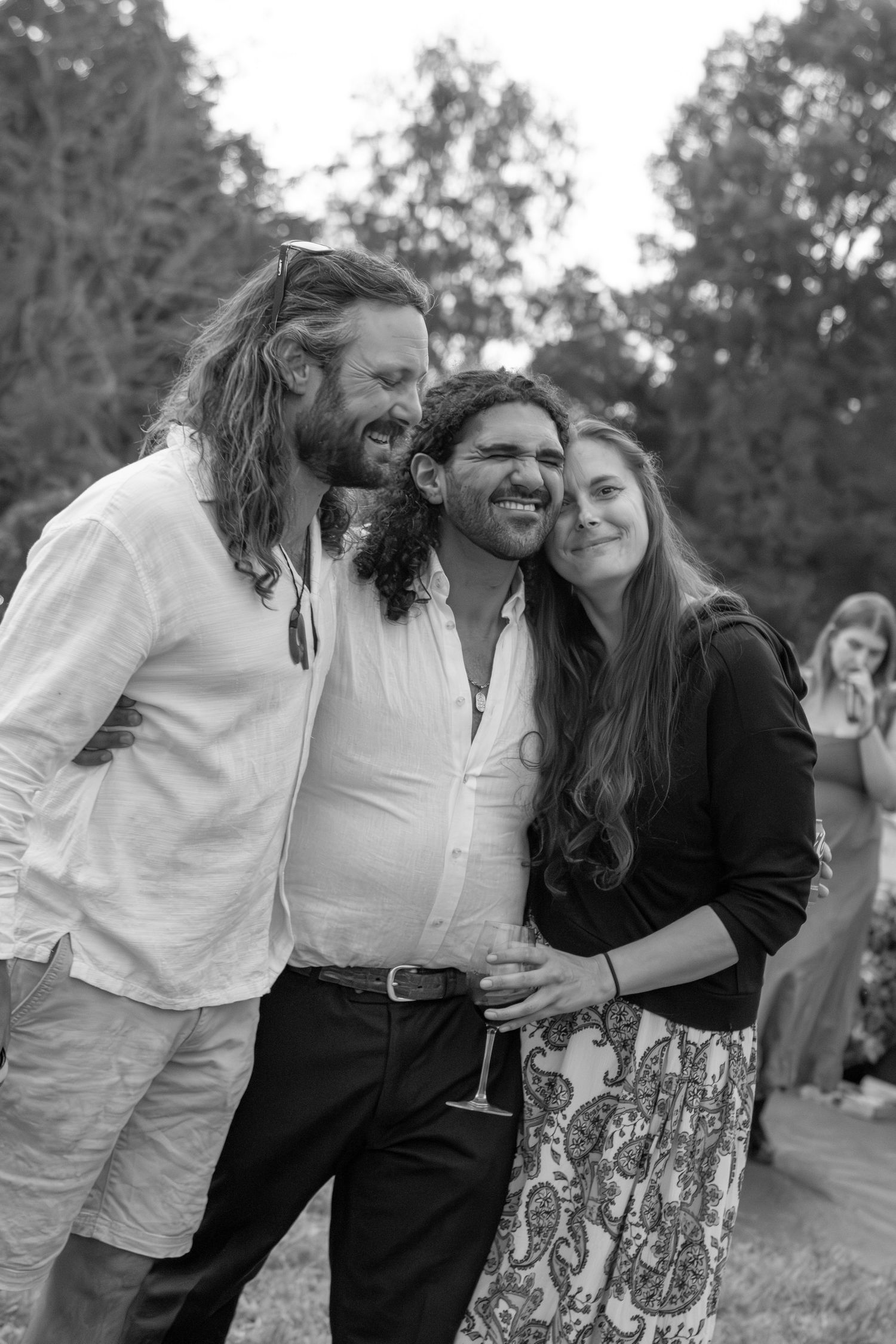 Black and white photo of three smiling friends posing together outdoors at a casual garden event.