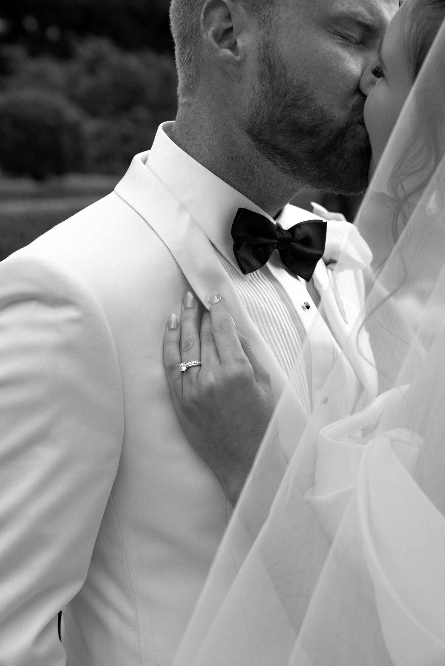 Black and white wedding photo of groom in white tuxedo and bow tie kissing bride, her hand resting on his chest.
