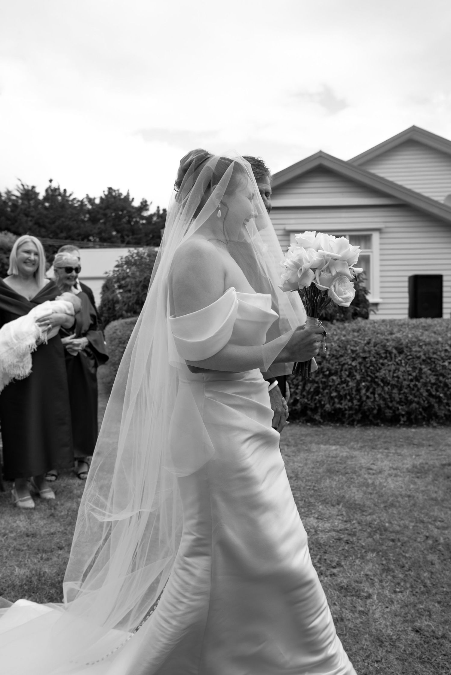 Bride in white off-shoulder gown and veil holds floral bouquet outdoors at wedding ceremony in black and white photo.