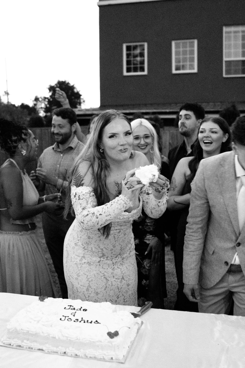 Bride cutting wedding cake at outdoor reception with guests watching, black and white photo.