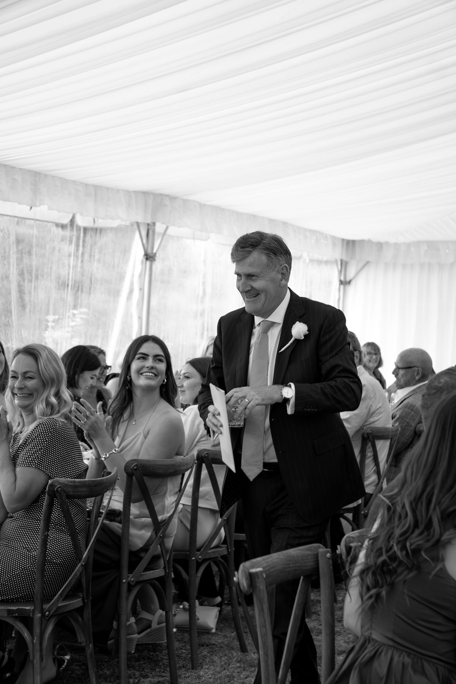 Black and white photo of a smiling man in a suit at a wedding reception inside a marquee tent.