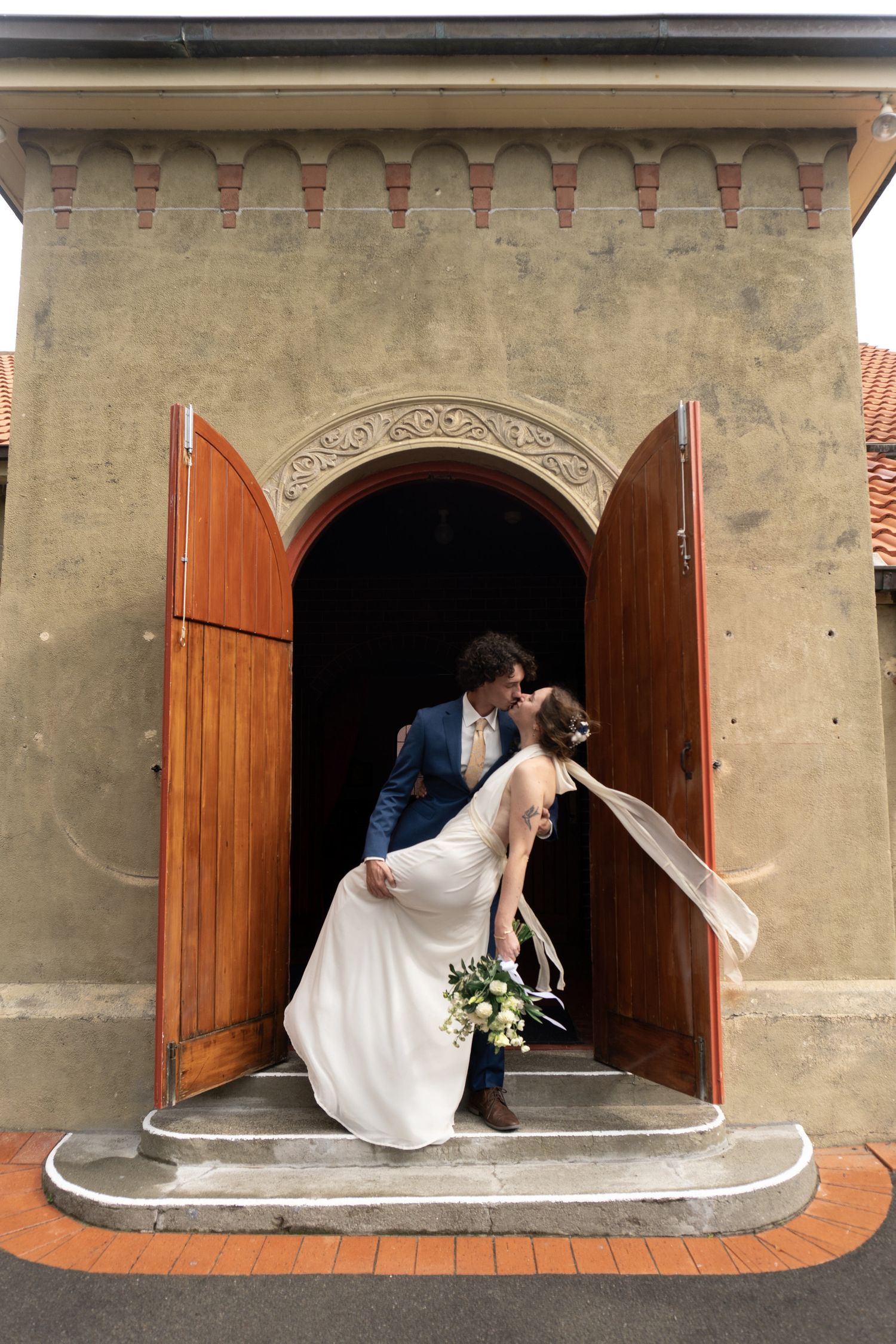 Bride and groom share a romantic kiss in an arched wooden doorway of a church, bride holding a bouquet.