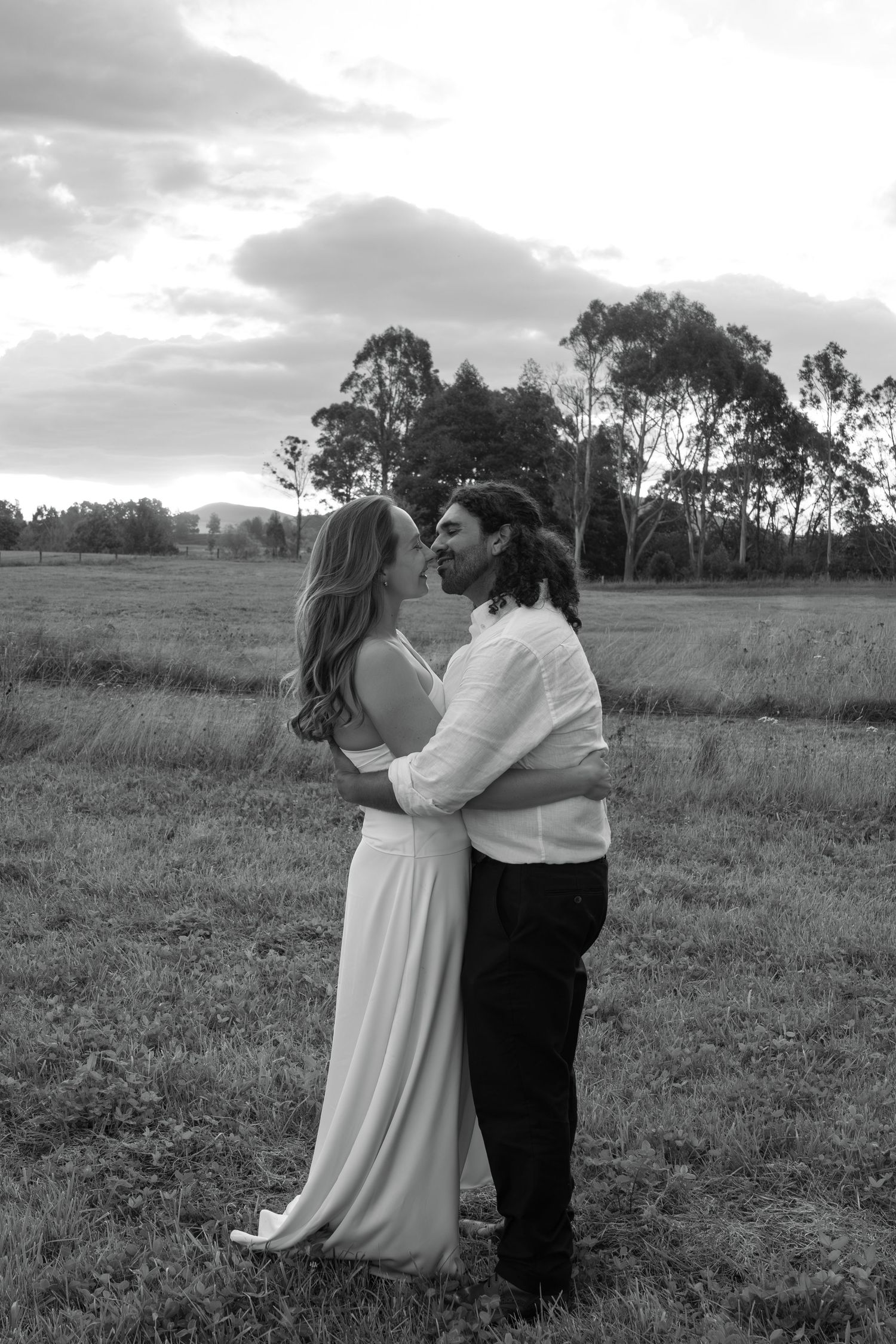 Black and white photo of a couple embracing in a field at sunset, woman in white dress, trees in background.