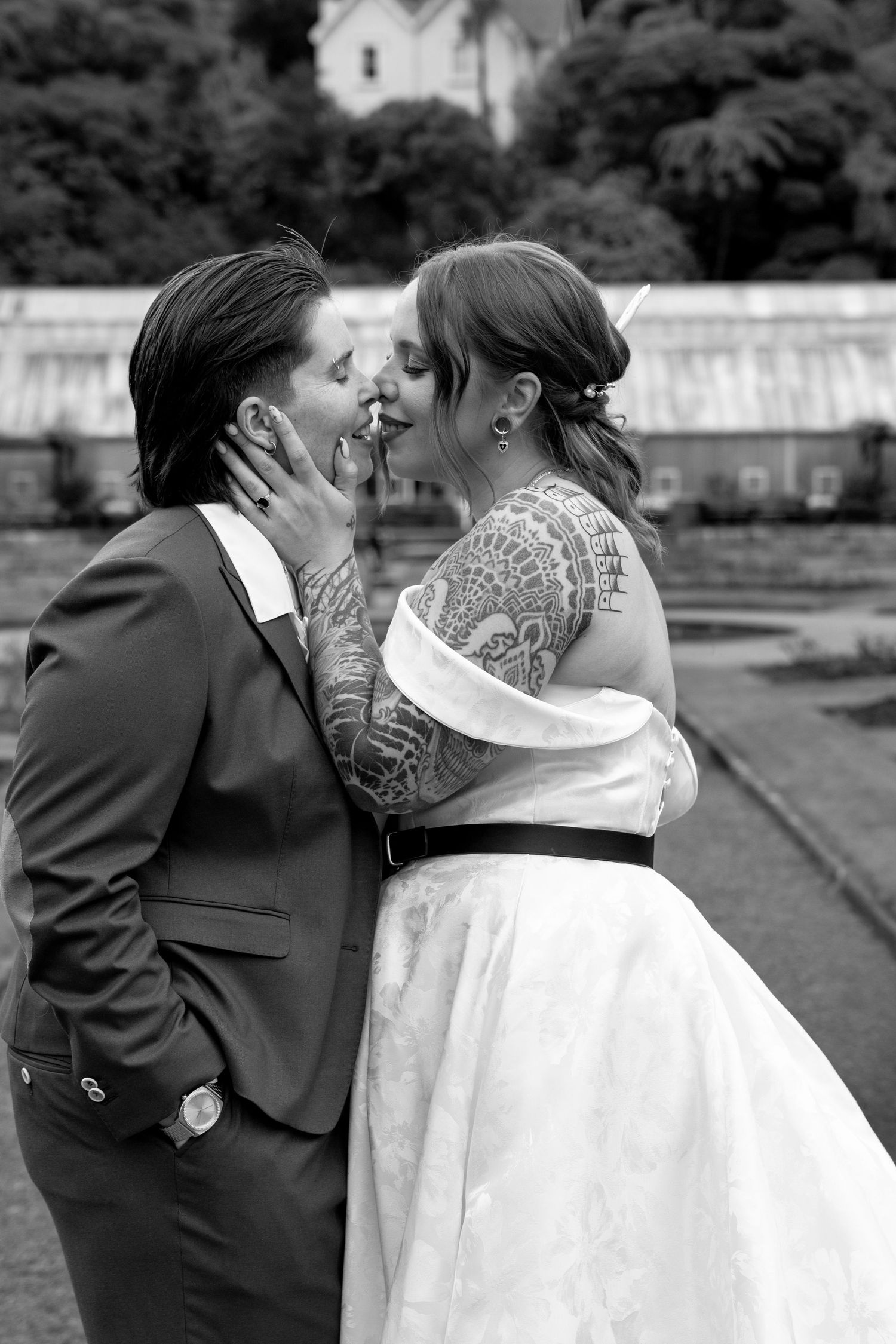 Black and white wedding photo of tattooed couple sharing a kiss, bride in white gown with black belt, groom in dark suit.