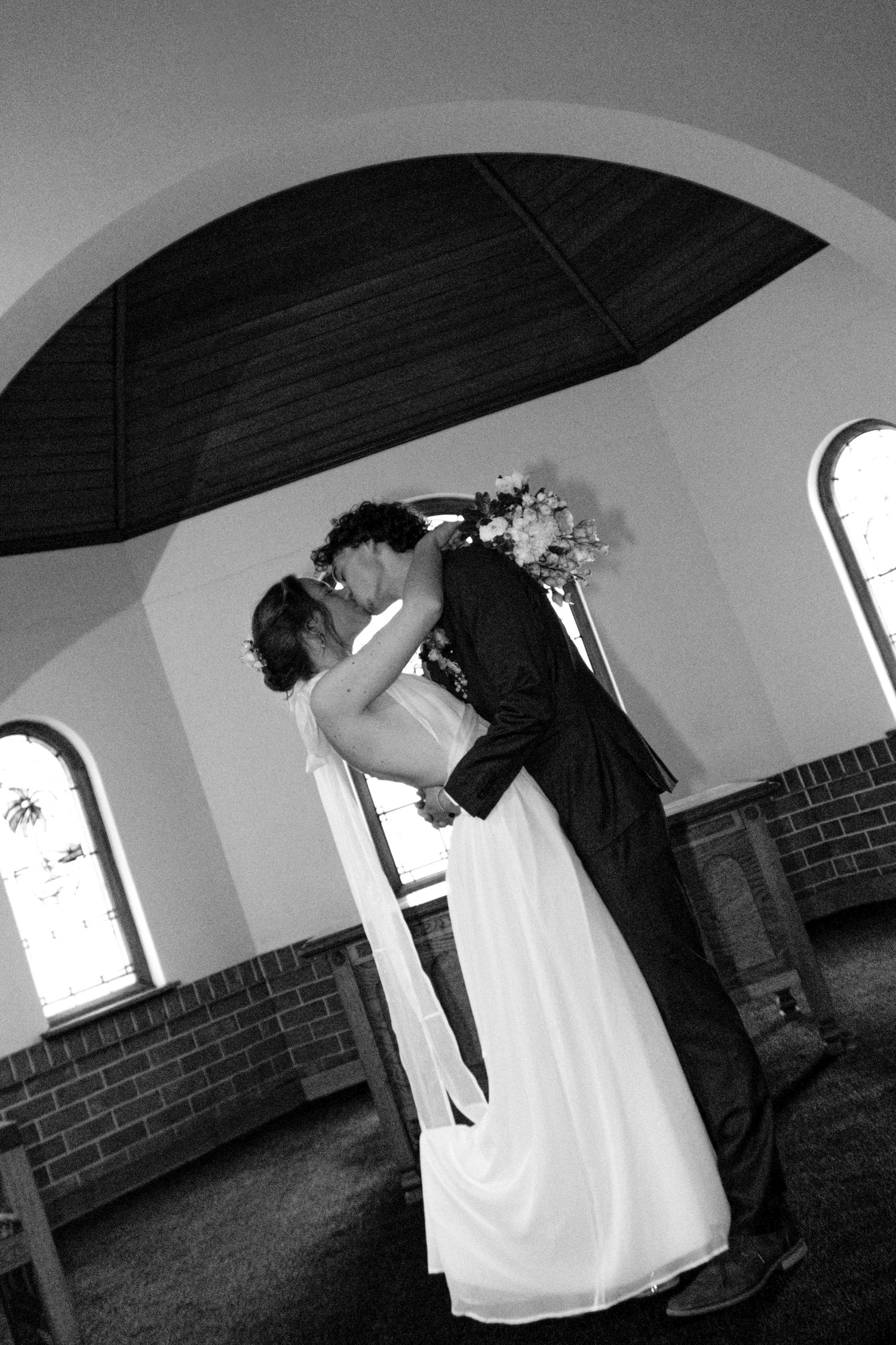 Black and white photo of bride and groom kissing under umbrella inside church on wedding day.