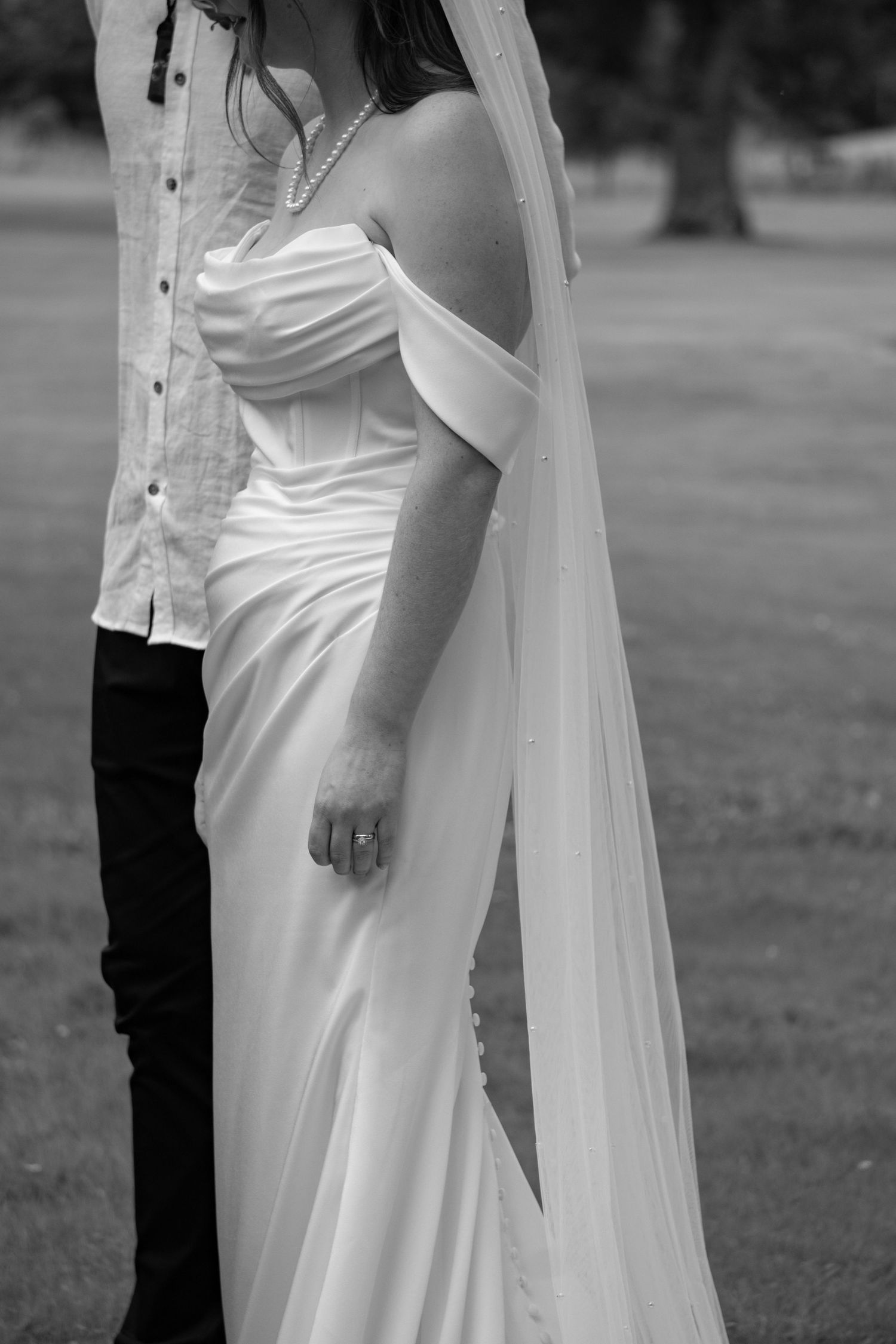 Black and white photo of a bride in an off-shoulder wedding gown with veil, standing beside a groom outdoors.