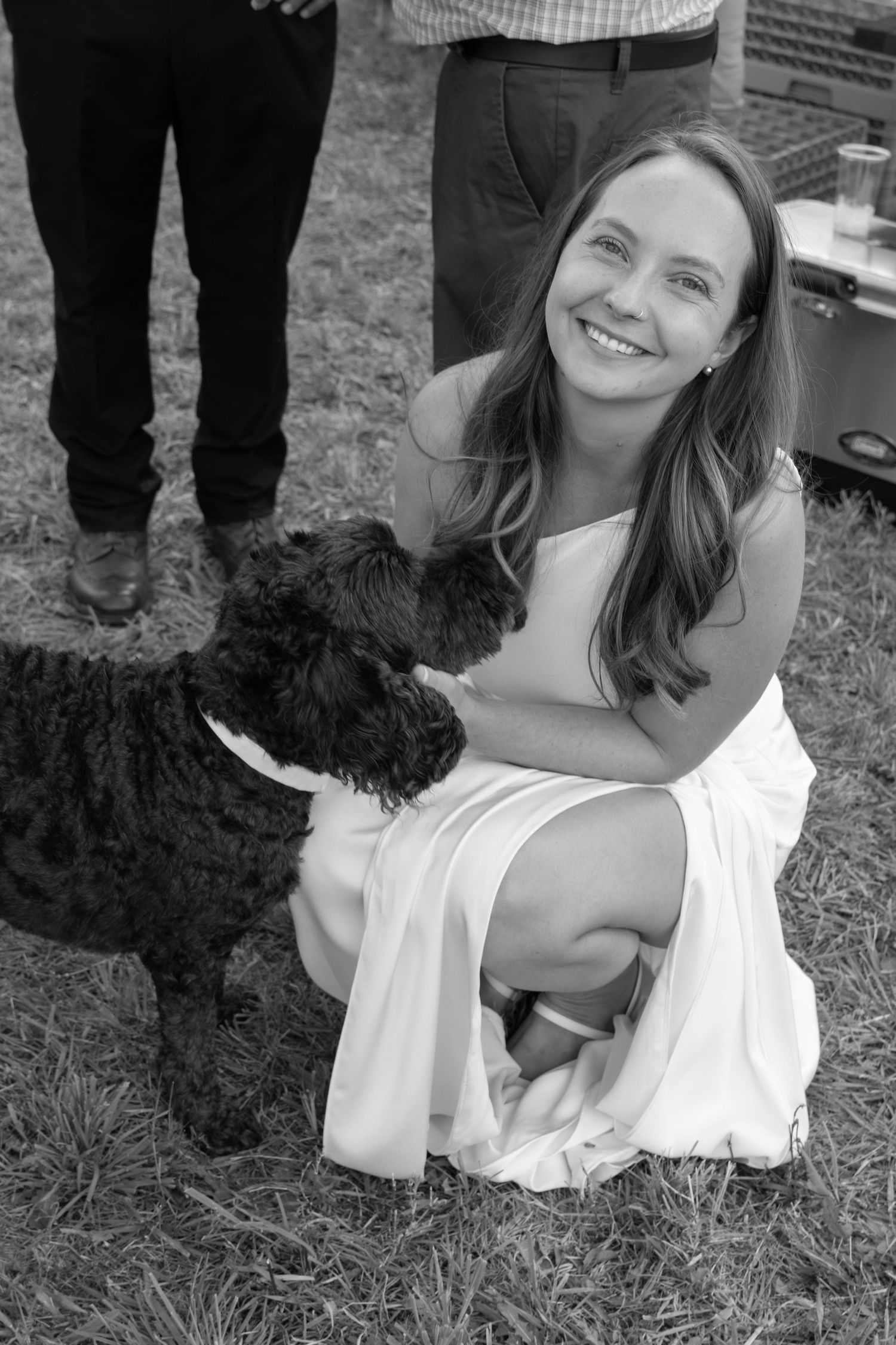 Black and white photo of a smiling woman in white dress crouching beside a curly black dog on grass at an outdoor event.