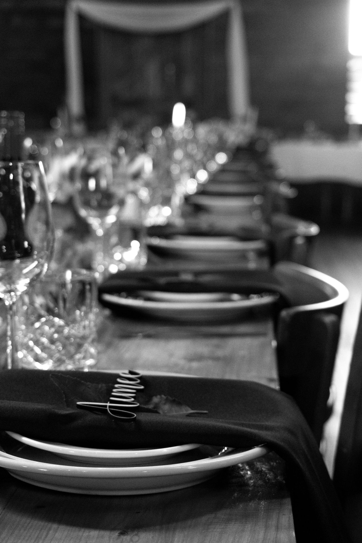 Black and white photo of an elegant long dining table set with stacked plates, dark napkins, and crystal glasses.