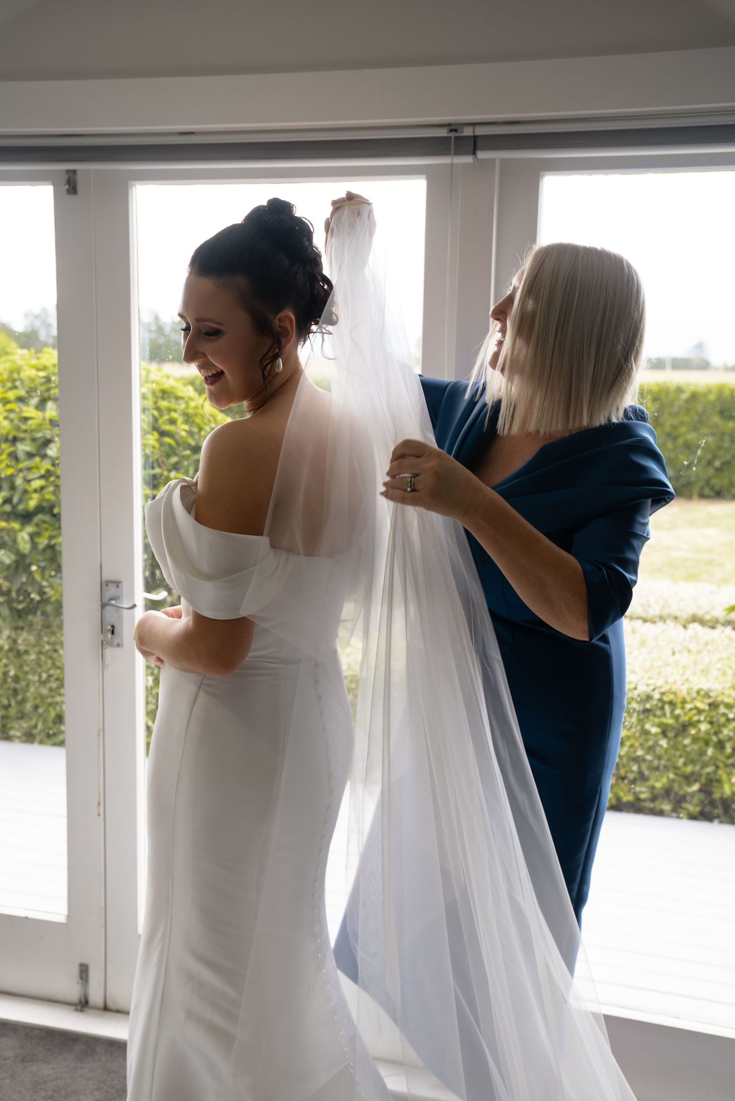 Bride in elegant white off-shoulder gown having veil adjusted by woman in blue dress indoors.
