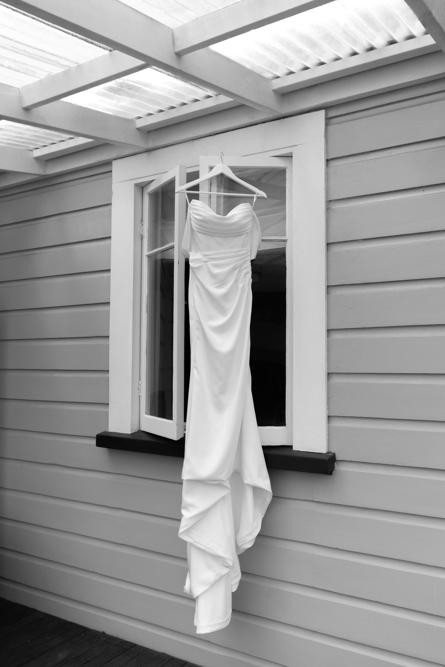 Black and white photo of an elegant white wedding dress hanging on a hanger outside a window of a weatherboard house.