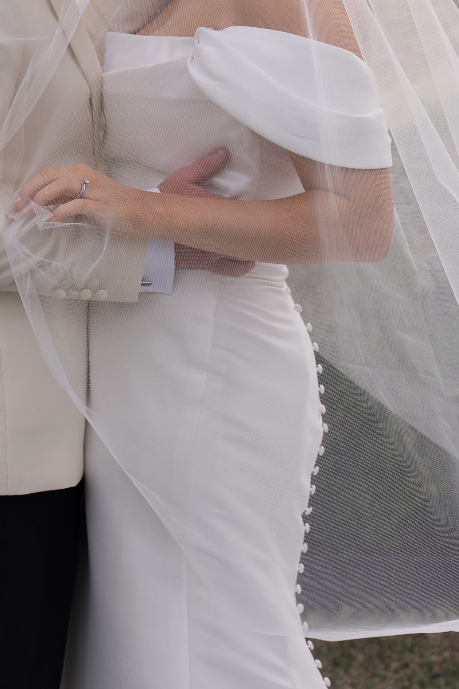 Bride in off-shoulder white gown with veil embracing groom in white tuxedo jacket at wedding ceremony.