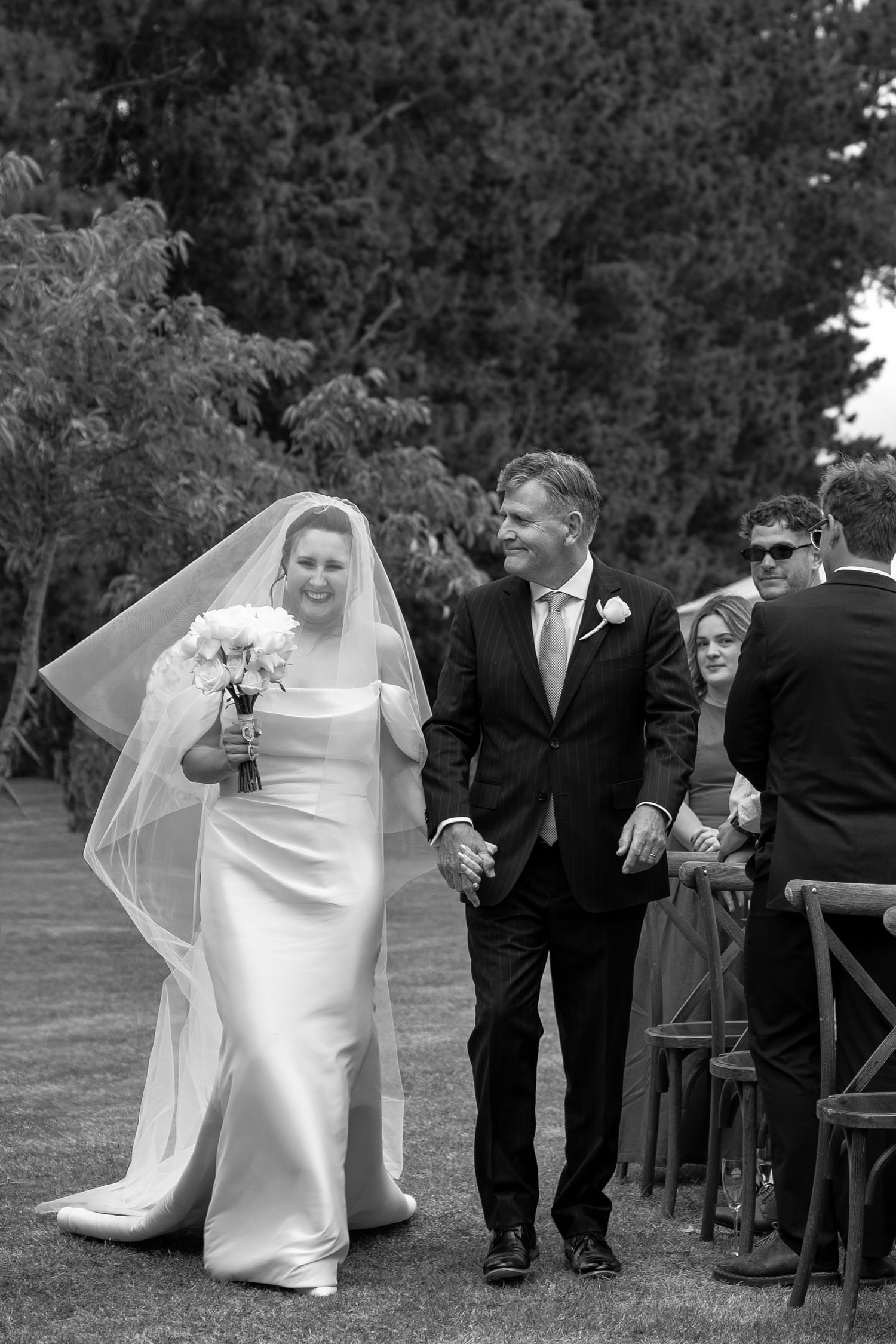 Black and white photo of a smiling bride in a veil holding flowers, walking the aisle with a man in a suit at an outdoor wedding.