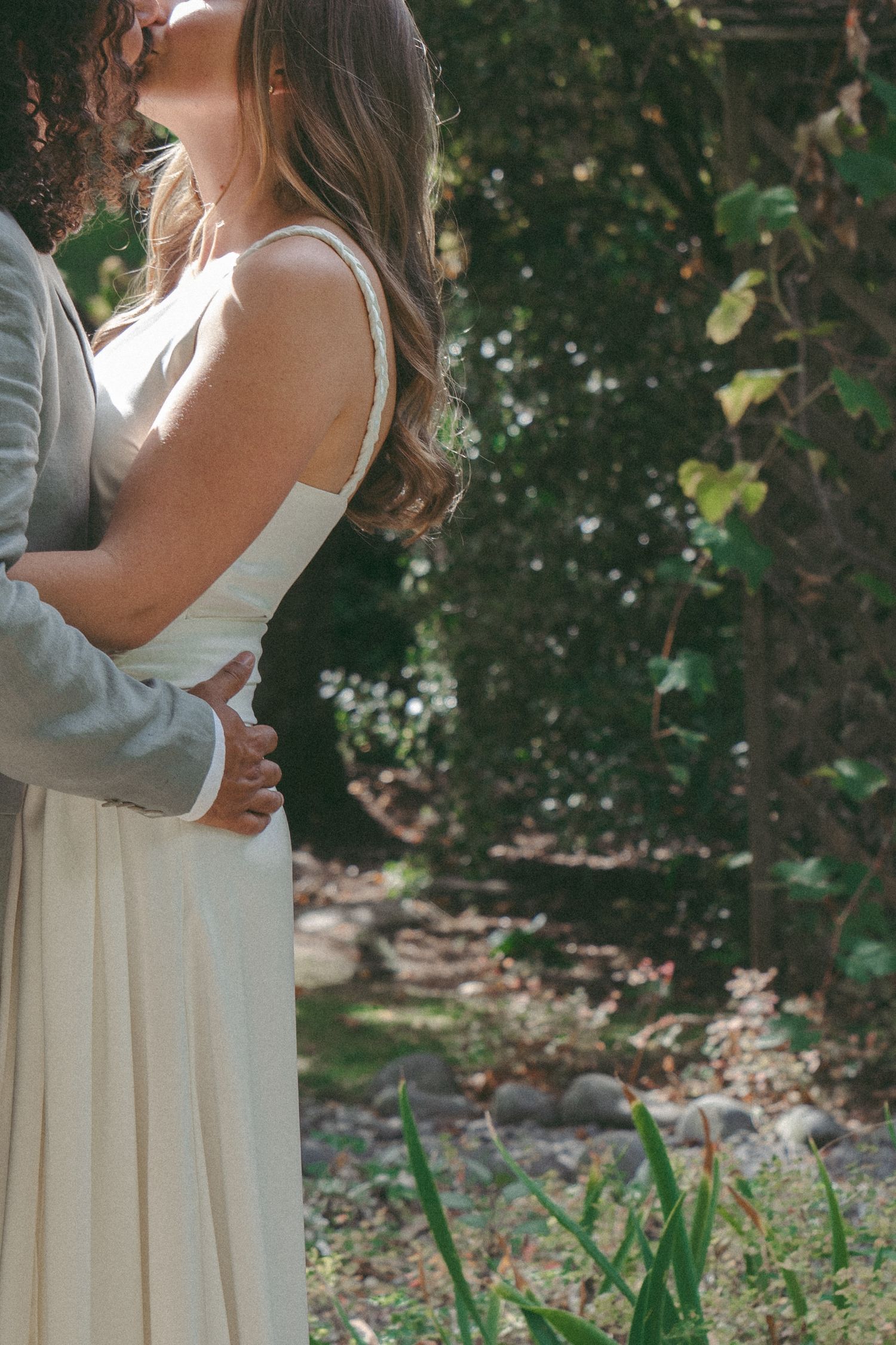 Couple embracing outdoors in garden setting, bride in white gown and groom in gray suit sharing romantic moment.