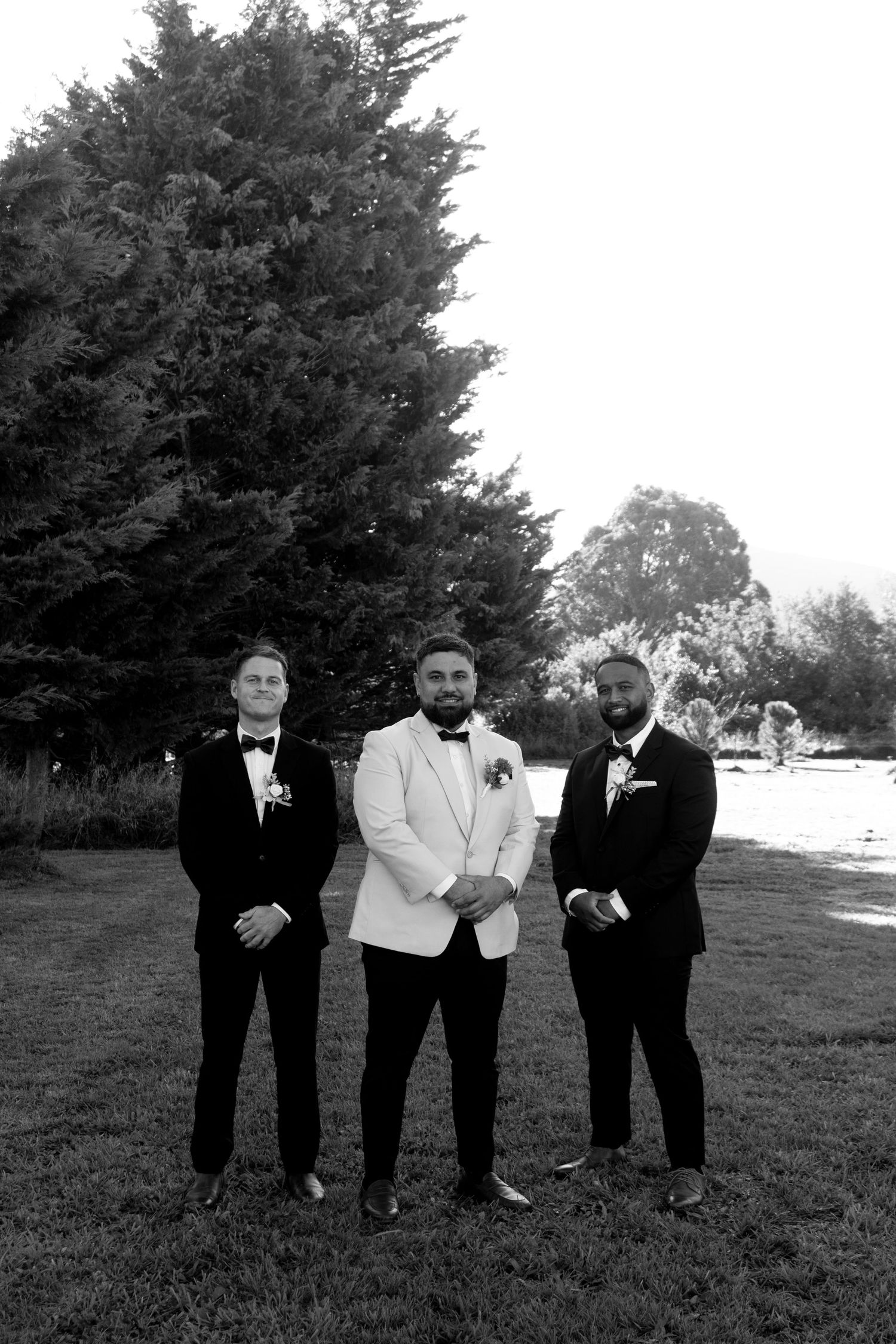 Black and white photo of groom in white tuxedo jacket flanked by two groomsmen in black tuxedos outdoors.