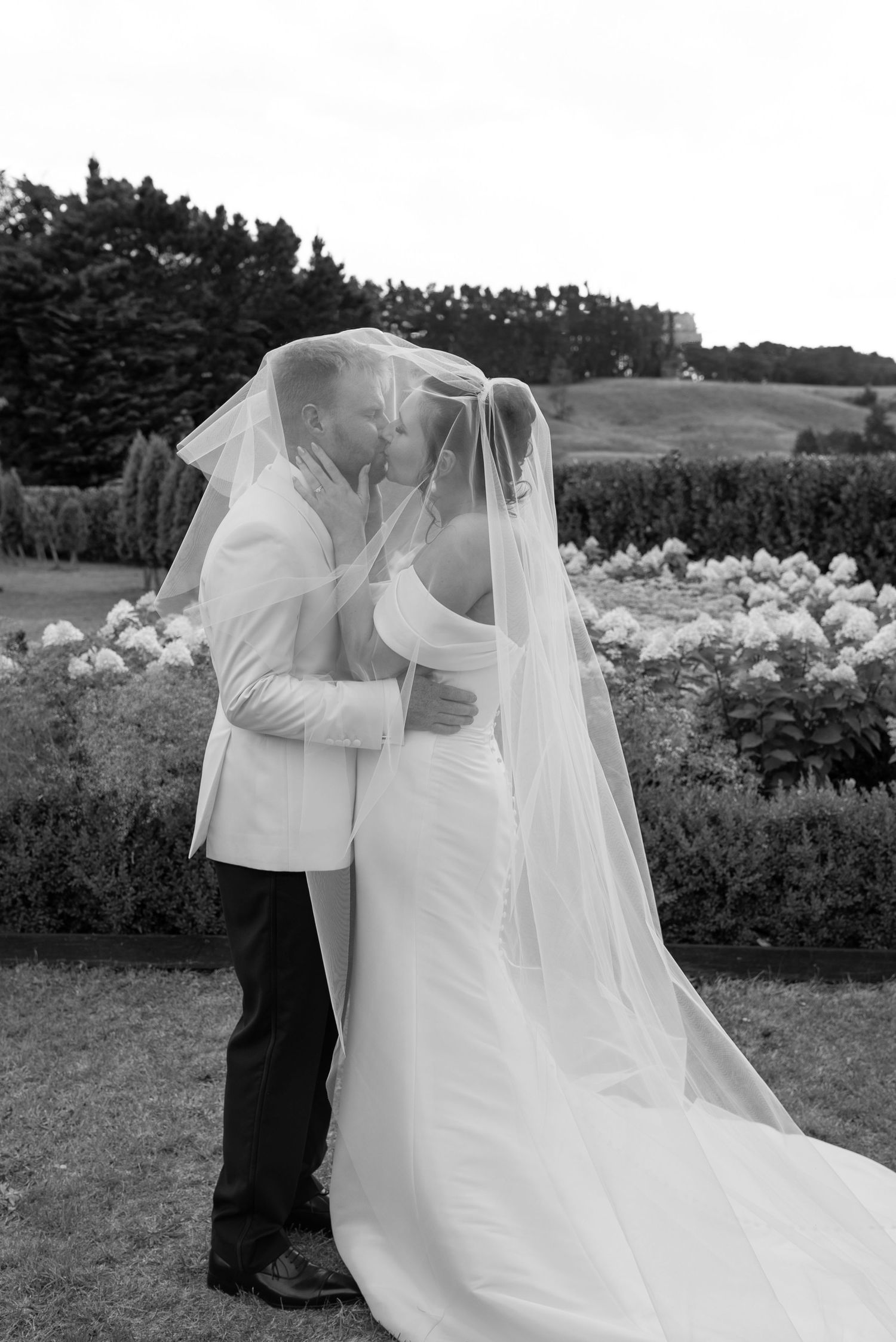 Black and white photo of a bride and groom kissing under a flowing veil in a garden setting.