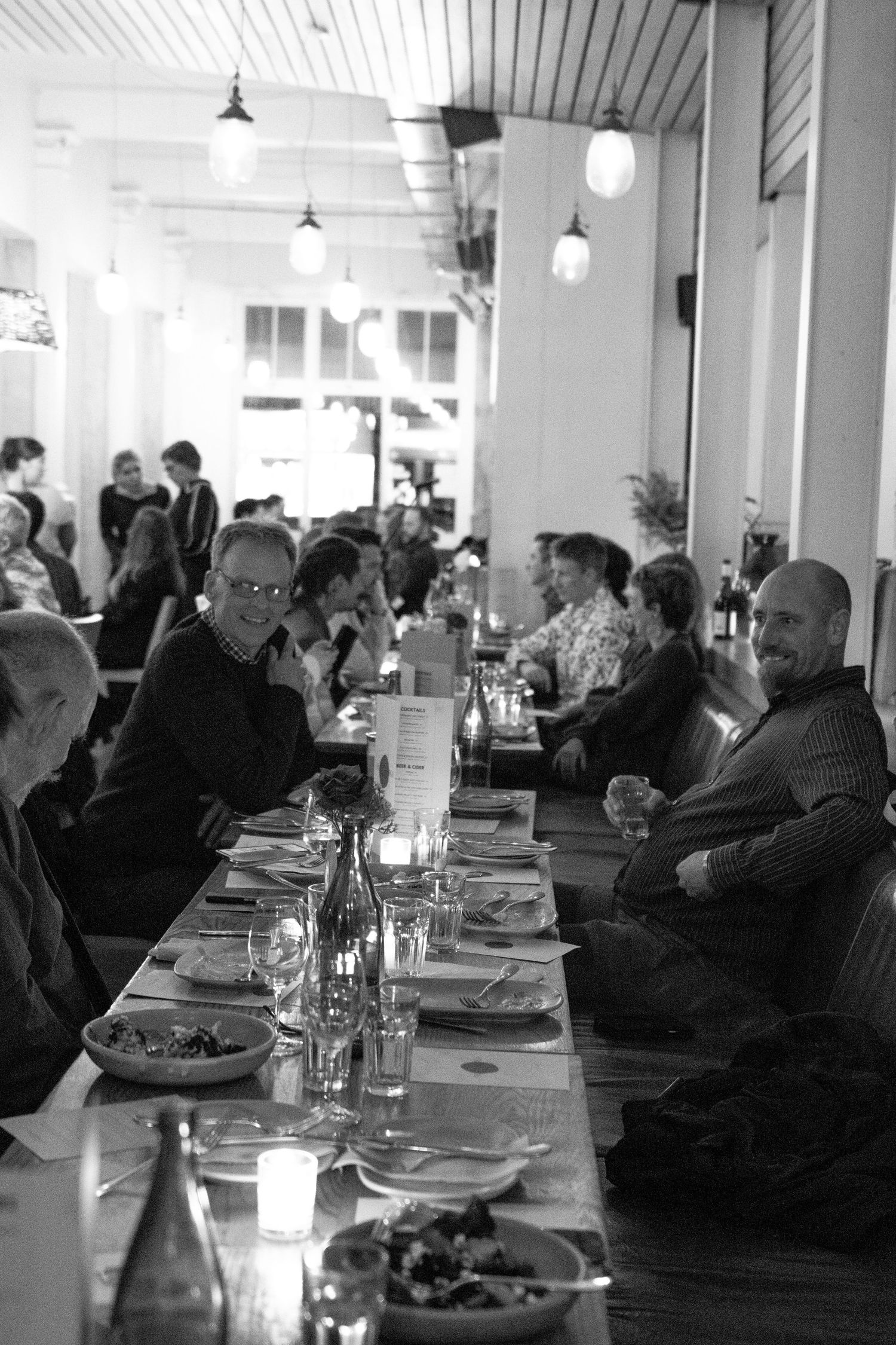 Black and white photo of guests dining at a long restaurant table with candles and bottles during an evening event.