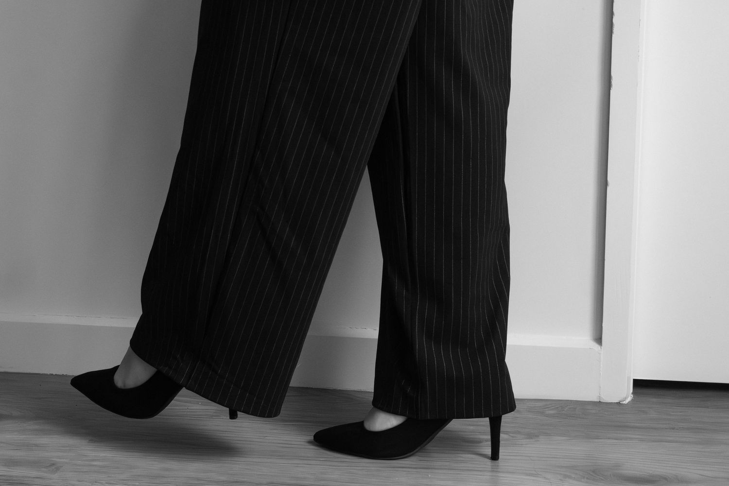 Black and white photo of woman wearing pinstripe wide-leg trousers and black stiletto heels standing by a door.