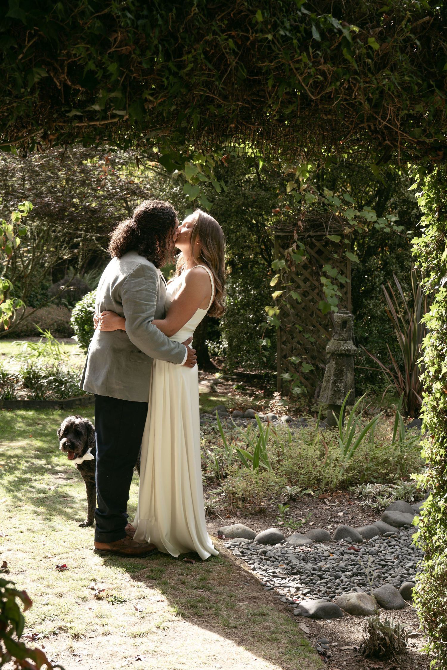 Bride and groom share a romantic kiss in a lush garden setting, with a small black dog nearby.