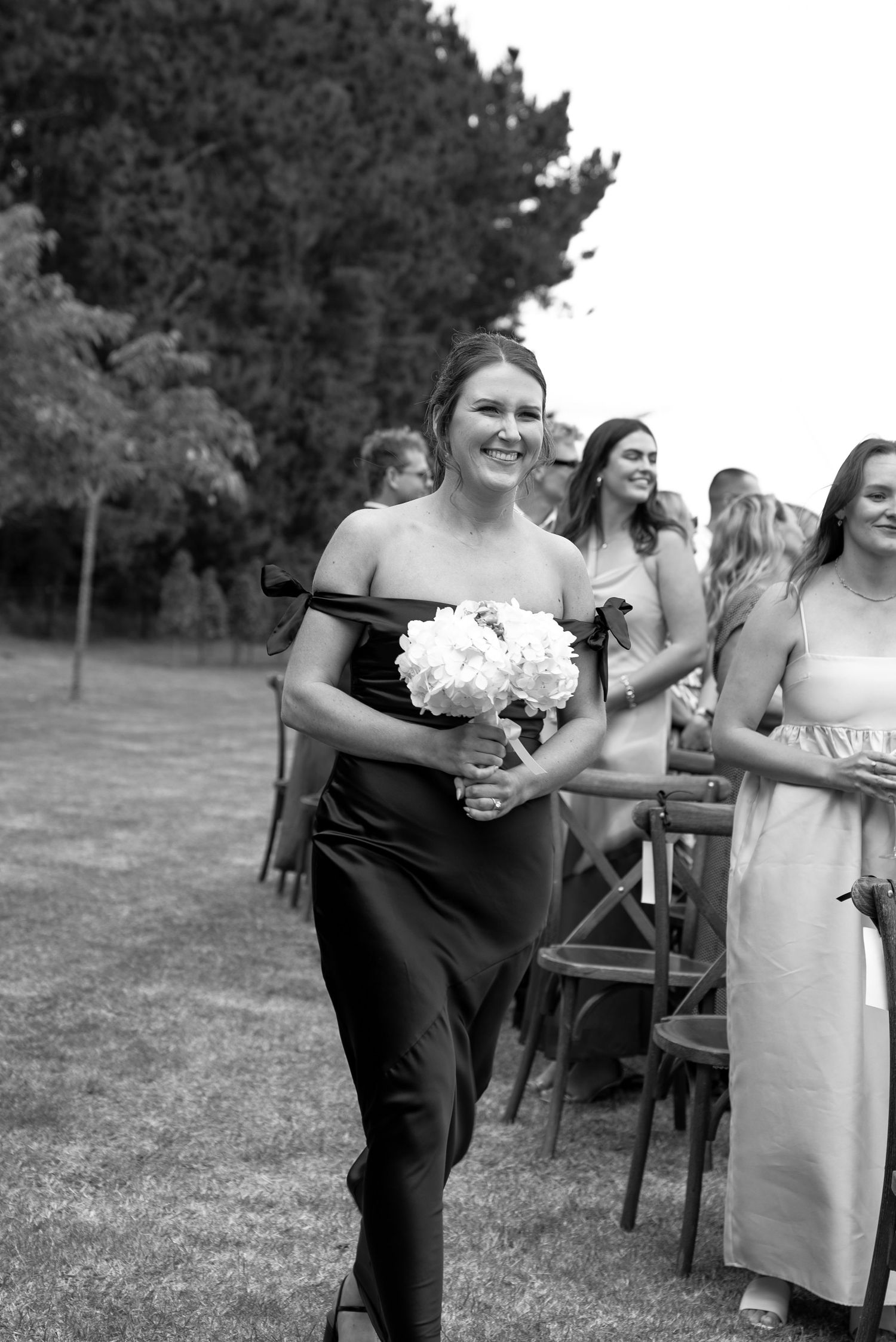 Smiling bridesmaid in black strapless gown walks down outdoor wedding aisle holding white peony bouquet.
